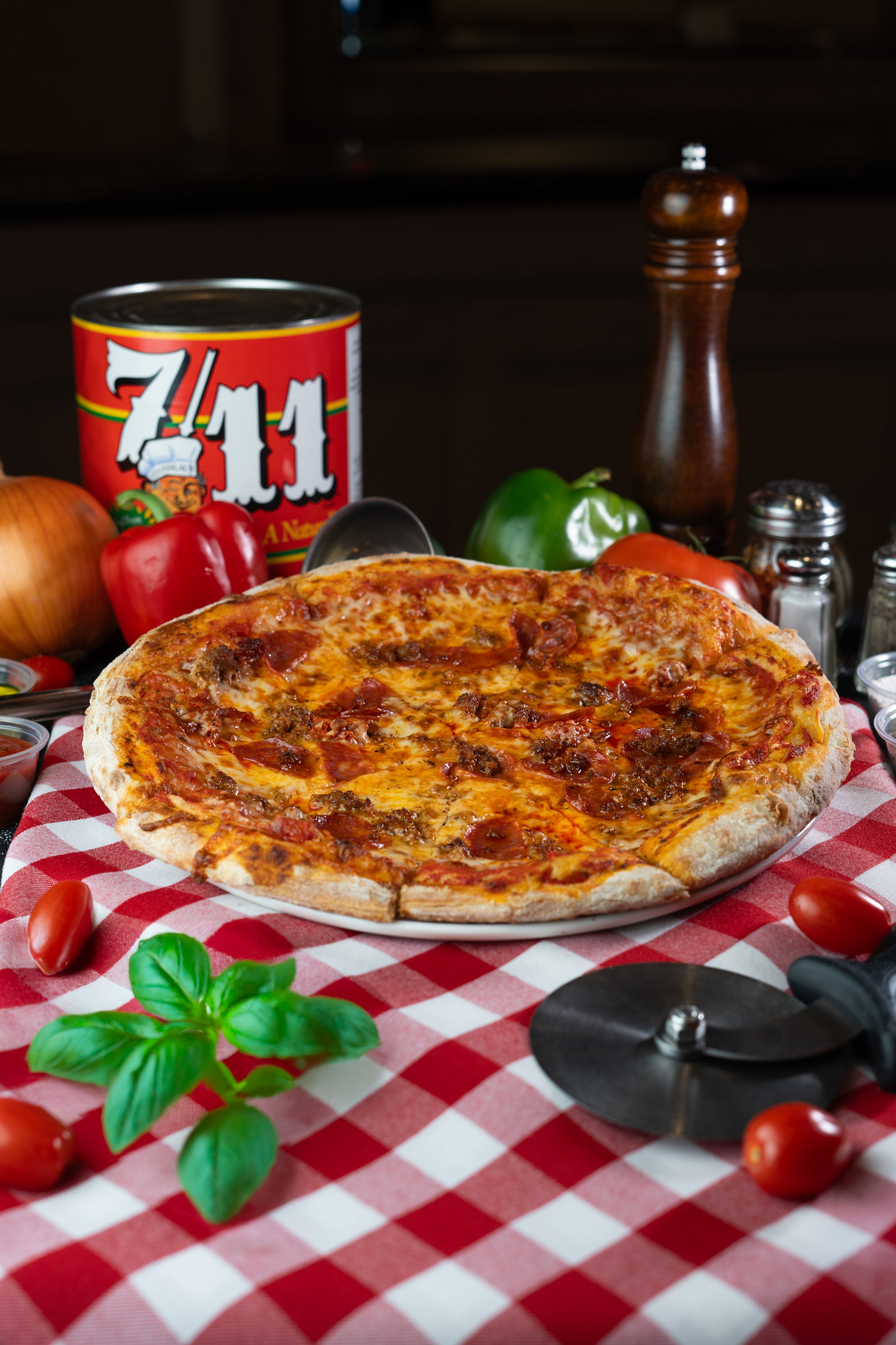 A pizza with pepperoni and sausage on a white plate, placed on a red and white checkered tablecloth, with fresh tomatoes, an onion, bell peppers, a basil plant, and various condiments and seasonings in the background.