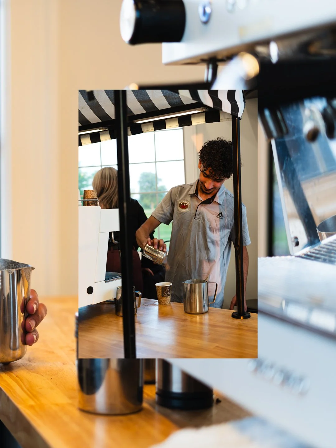 Barista pouring milk into a cup at a coffee shop with a striped awning and large windows in the background.
