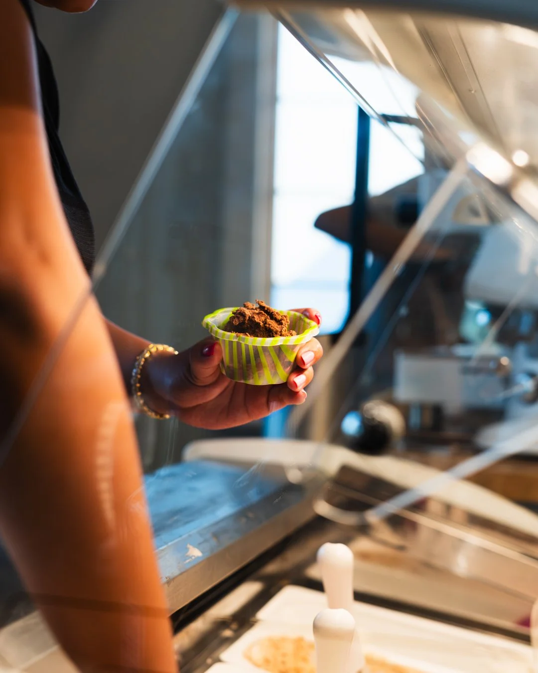 Person holding a green and yellow striped cup of chocolate ice cream at an ice cream shop, with a metal counter and ice cream toppings visible.