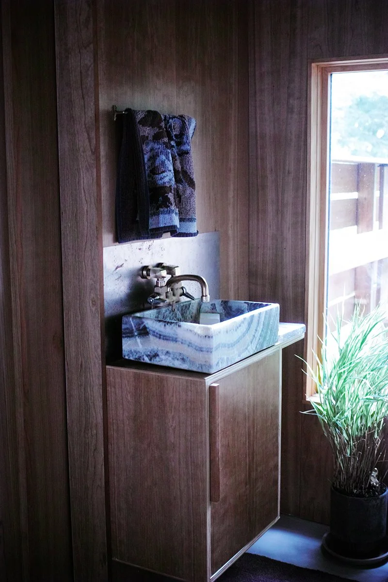 Wood-paneled wall with a stone sink, a towel hanging above, a window allowing natural light, and a potted plant on the floor.