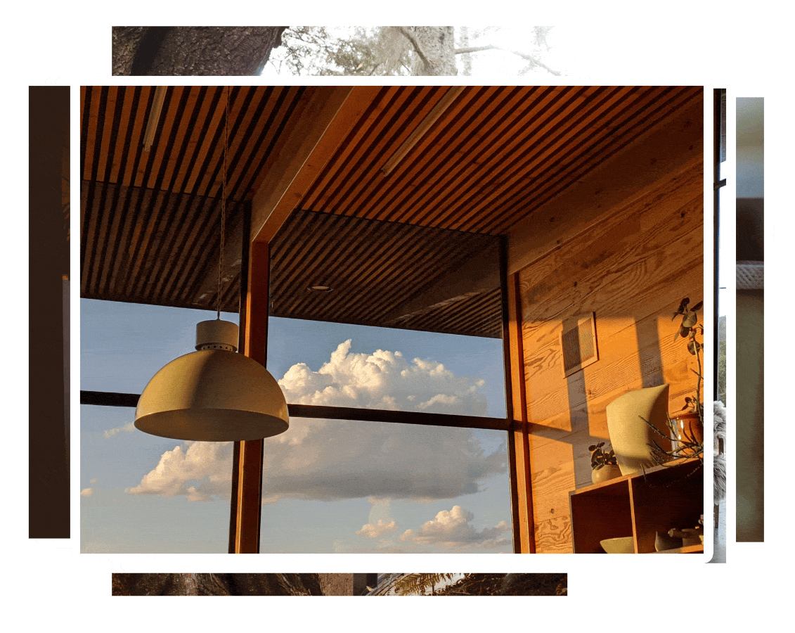 Interior view of a modern room with wooden walls and ceiling, large window showing a partly cloudy sky, hanging beige pendant lamp, and decorative plants on a shelf.