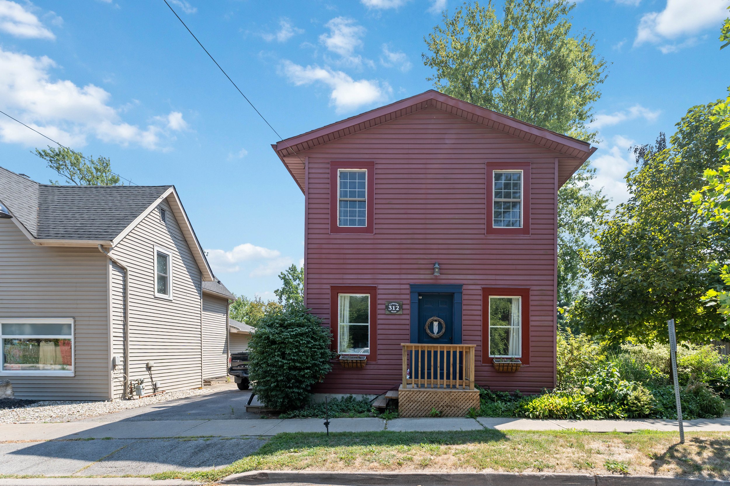 A two-story pink house with a blue door and three windows, surrounded by greenery under a partly cloudy sky.
