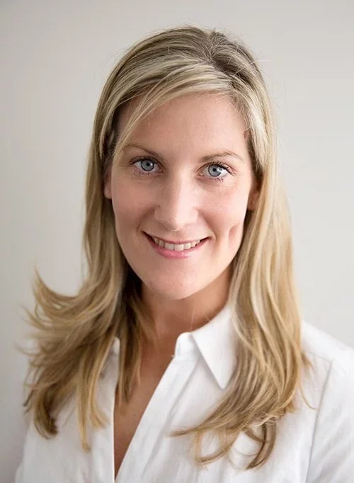 A blonde woman with blue eyes wearing a white blouse and smiling at the camera.