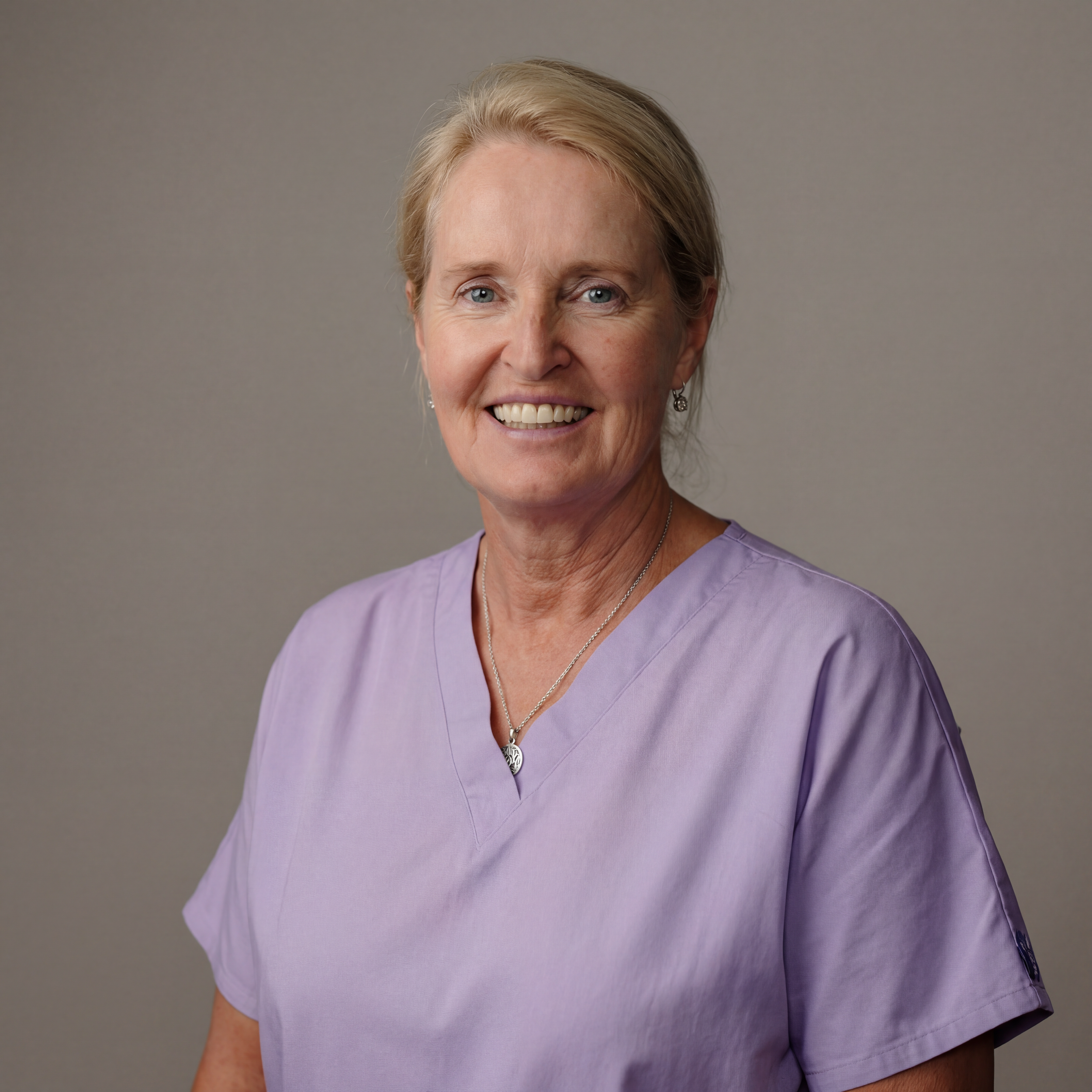 A middle-aged woman with blonde hair, smiling, wearing a light purple medical scrubs top, silver jewelry, and standing against a plain gray background.