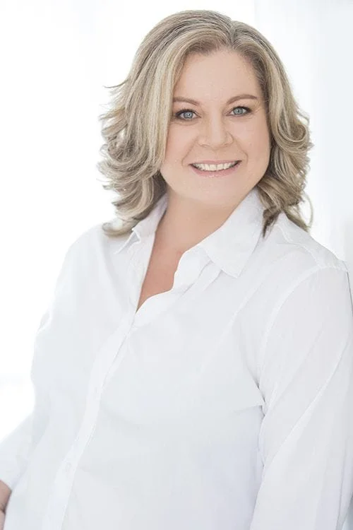 A woman with shoulder-length blonde hair, blue eyes, and a bright smile, wearing a white button-up shirt against a plain white background.