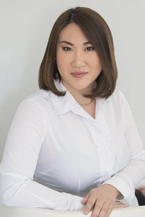 A woman with shoulder-length brown hair and light makeup, wearing a white button-up shirt, sitting at a desk with a neutral background.