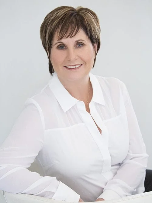 A woman with short brown hair, wearing a white blouse, smiling at the camera against a plain background.