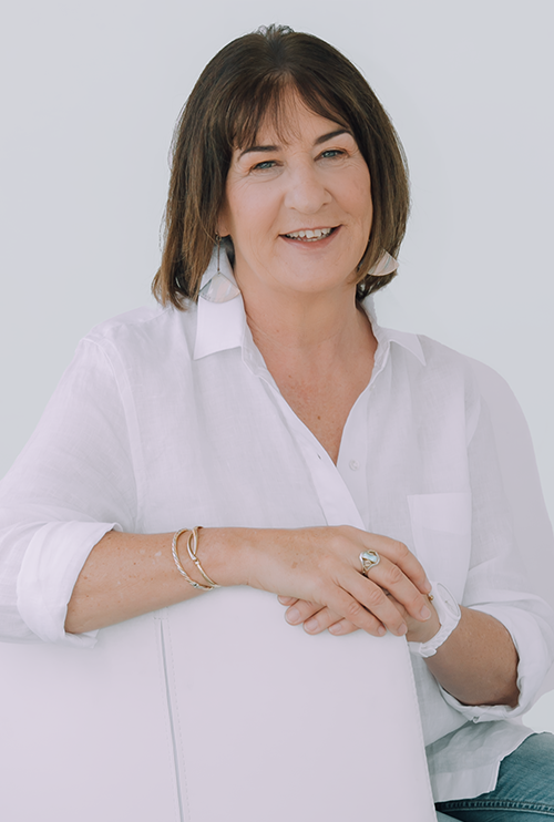 A middle-aged woman with shoulder-length brown hair and light skin, smiling and wearing a white button-up shirt with rolled-up sleeves, is sitting in front of a plain light background.