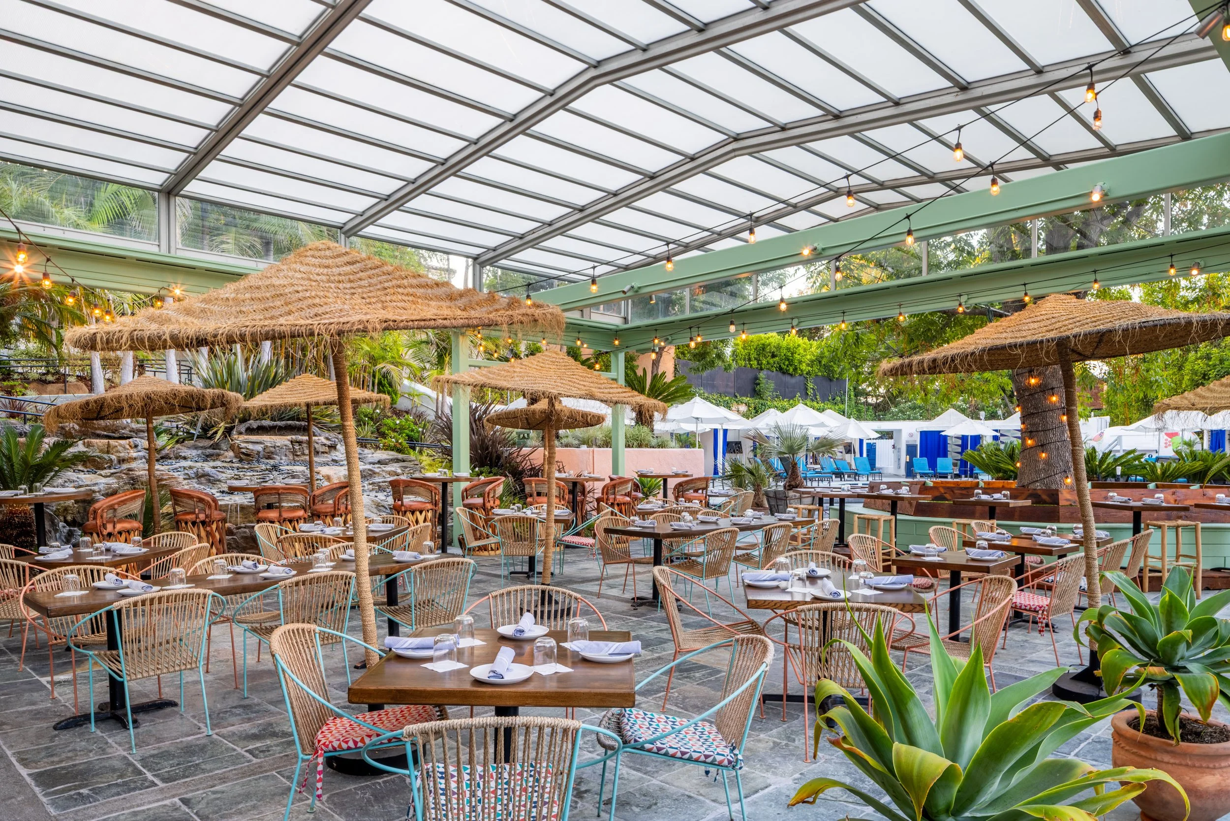 Outdoor restaurant patio with tables set for dining, surrounded by lush greenery and tropical plants, under a glass ceiling with string lights and straw umbrellas.