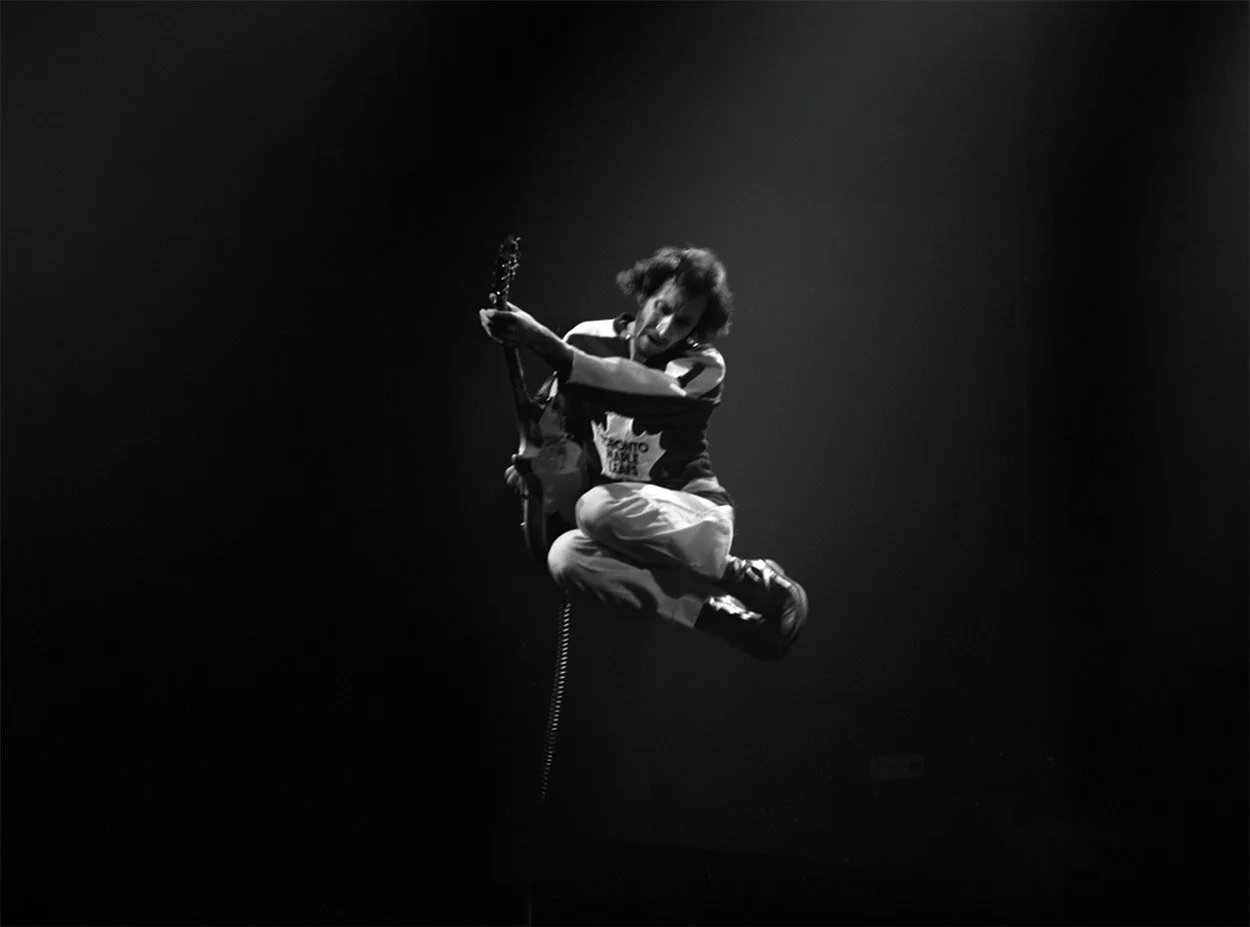A person jumping in the air while playing an electric guitar, captured in black and white with a dark background.