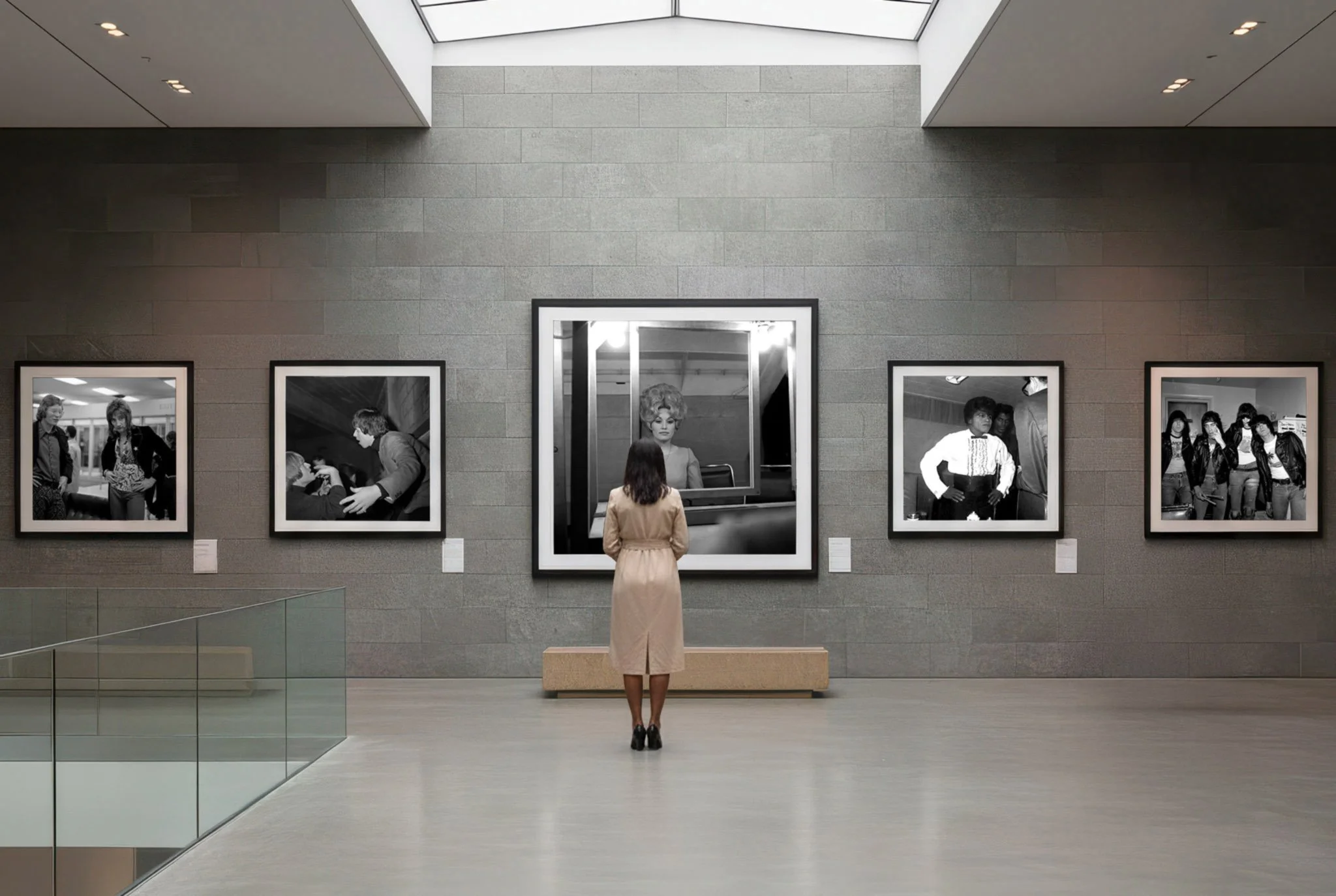 A woman stands in an art gallery looking at black-and-white photographs on a gray wall, with a large mirror reflecting her face.