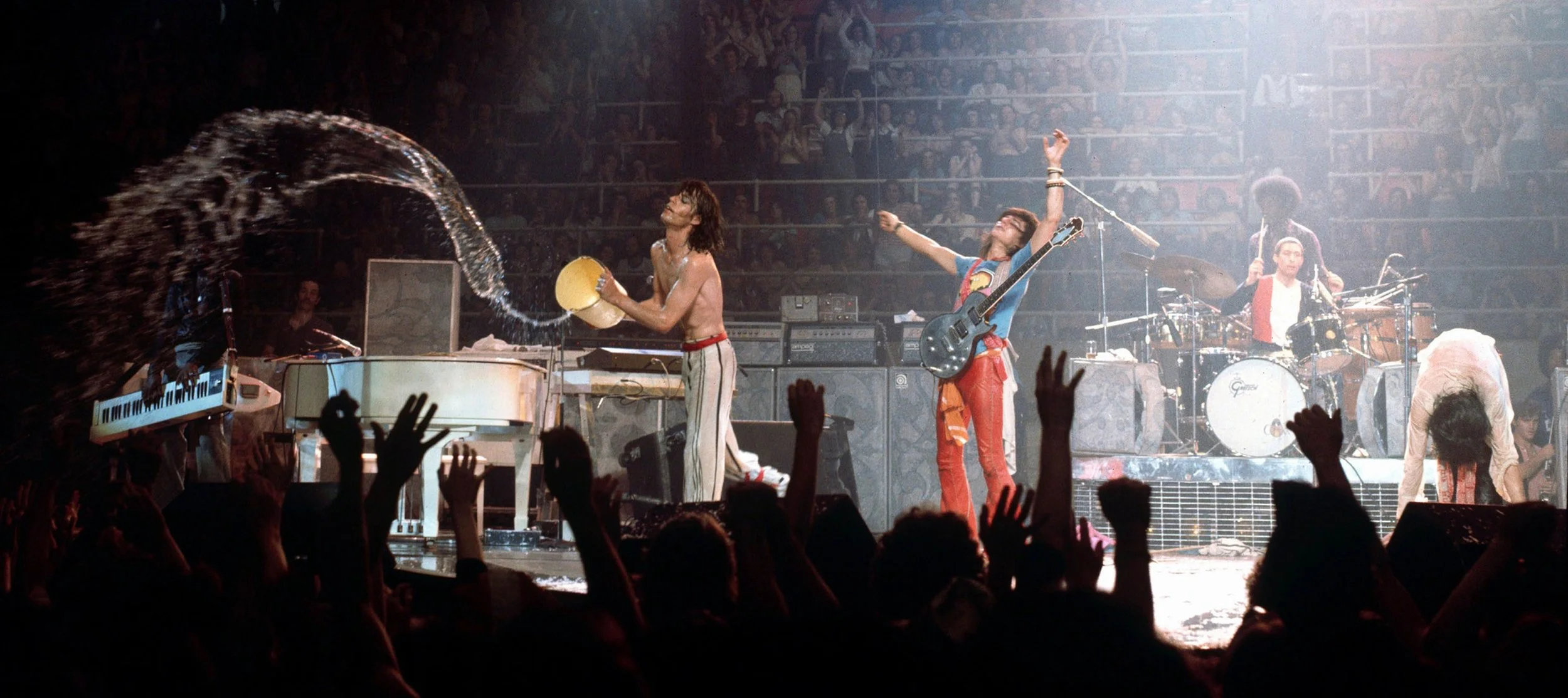 Band performing on stage with audience in front, one musician pouring water over a piano, others playing instruments, and stage lights illuminating the scene.