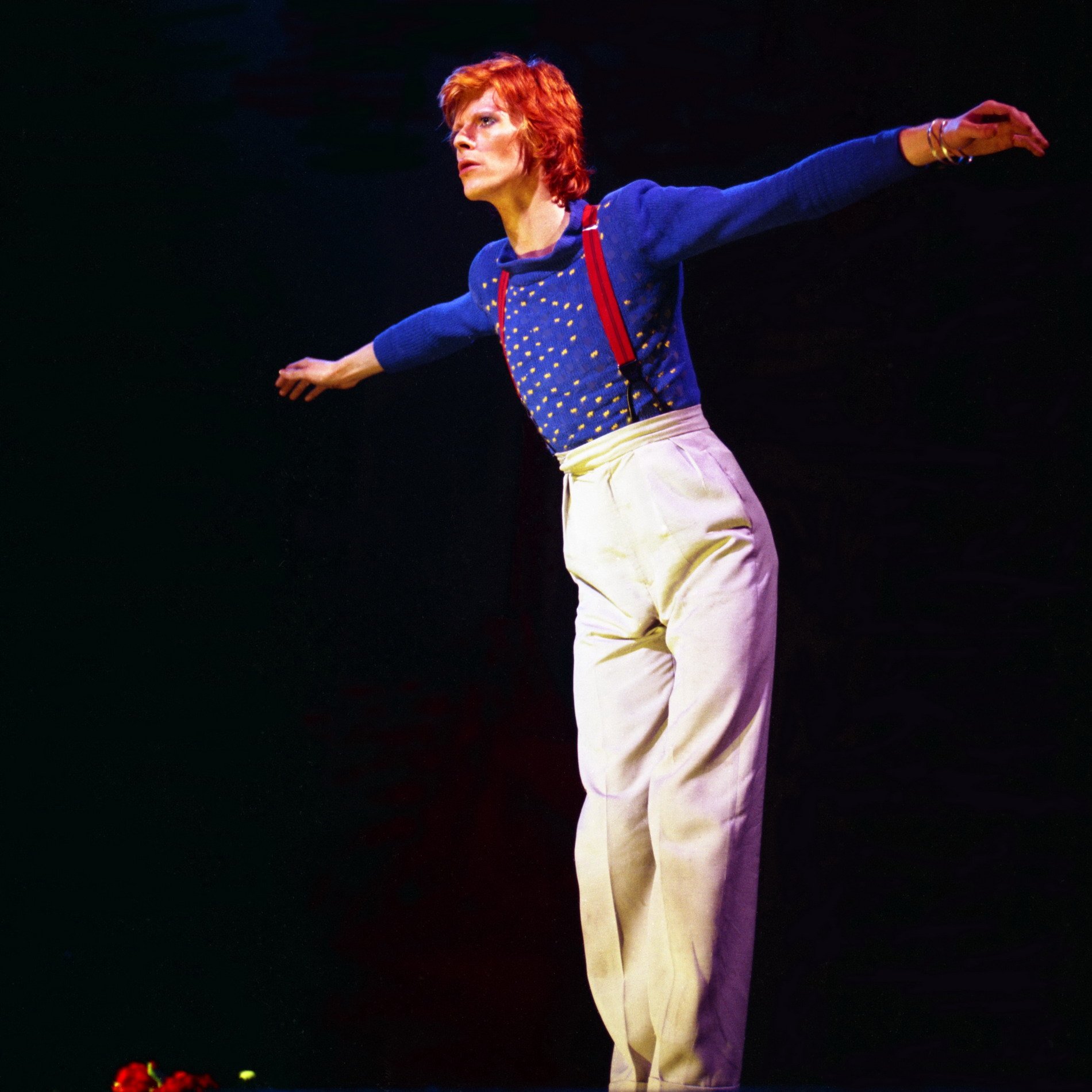 A woman with red hair performing on stage, wearing a blue sweater with yellow dots and white high-waisted pants, with her arms extended outward.