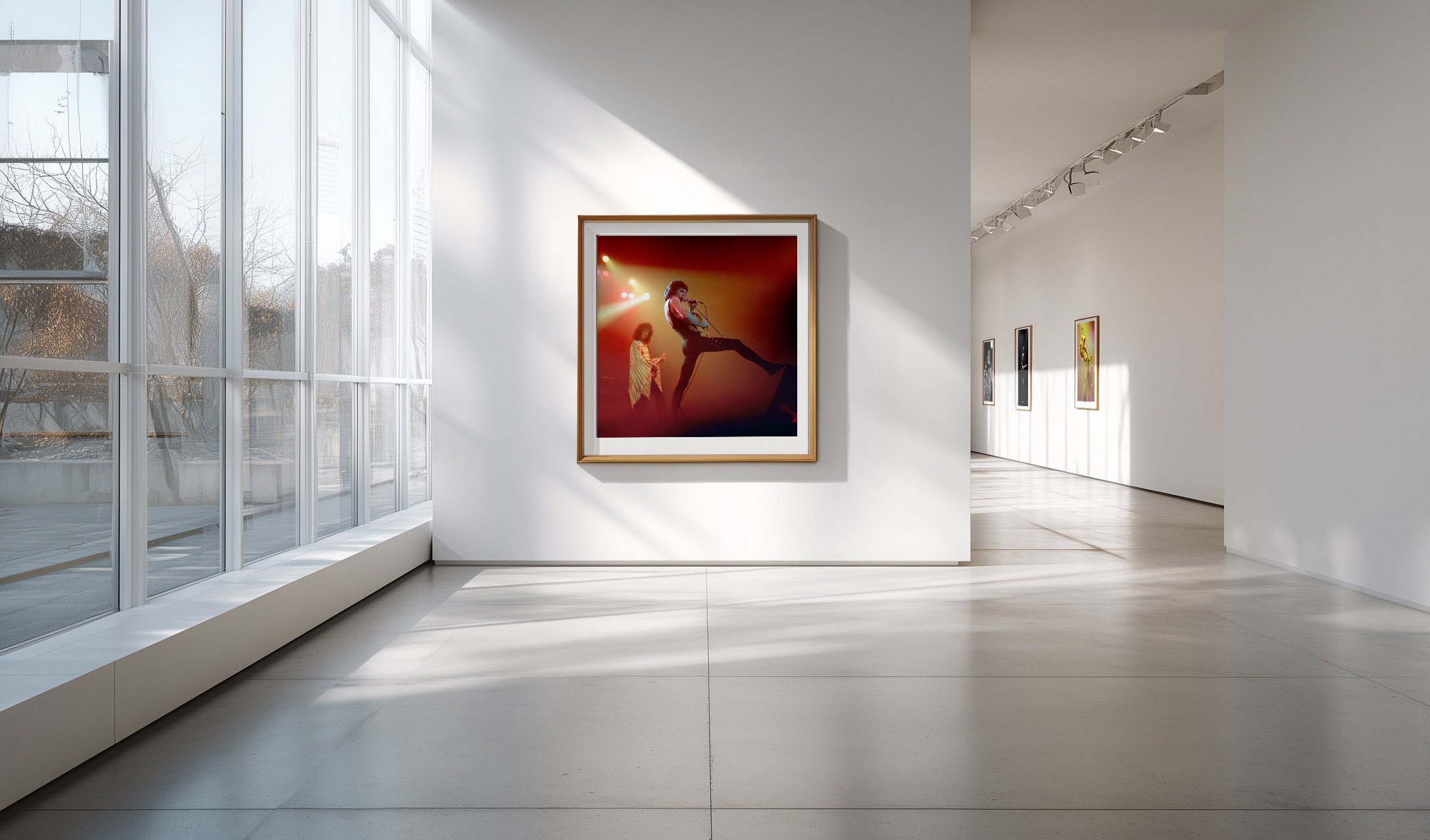 A modern art gallery with white walls and a large window on the left, displaying framed artwork including a photo of two performers on stage, one playing guitar and the other singing, illuminated with red and yellow lighting.