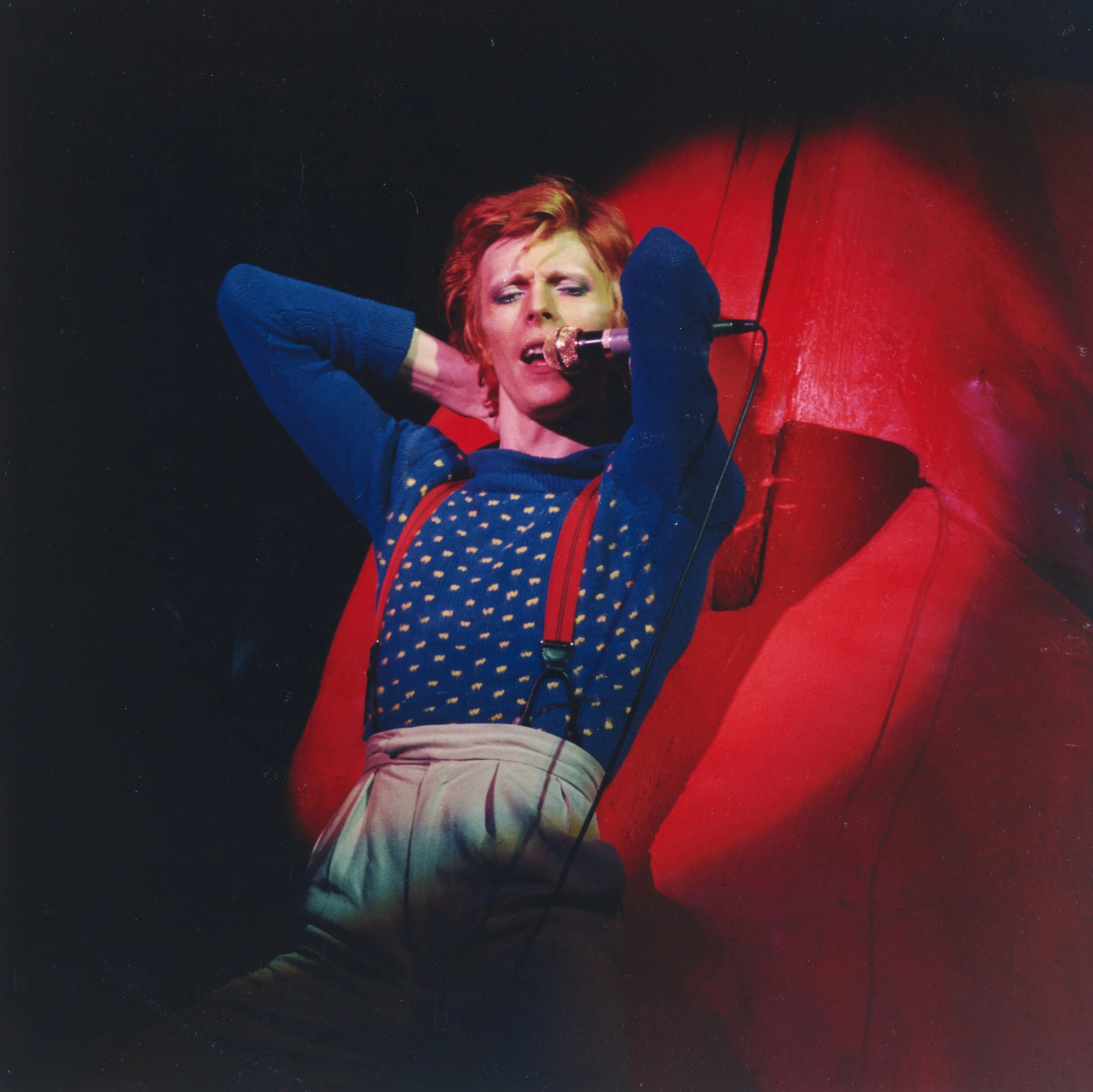 A woman with red hair singing into a microphone against a red background.