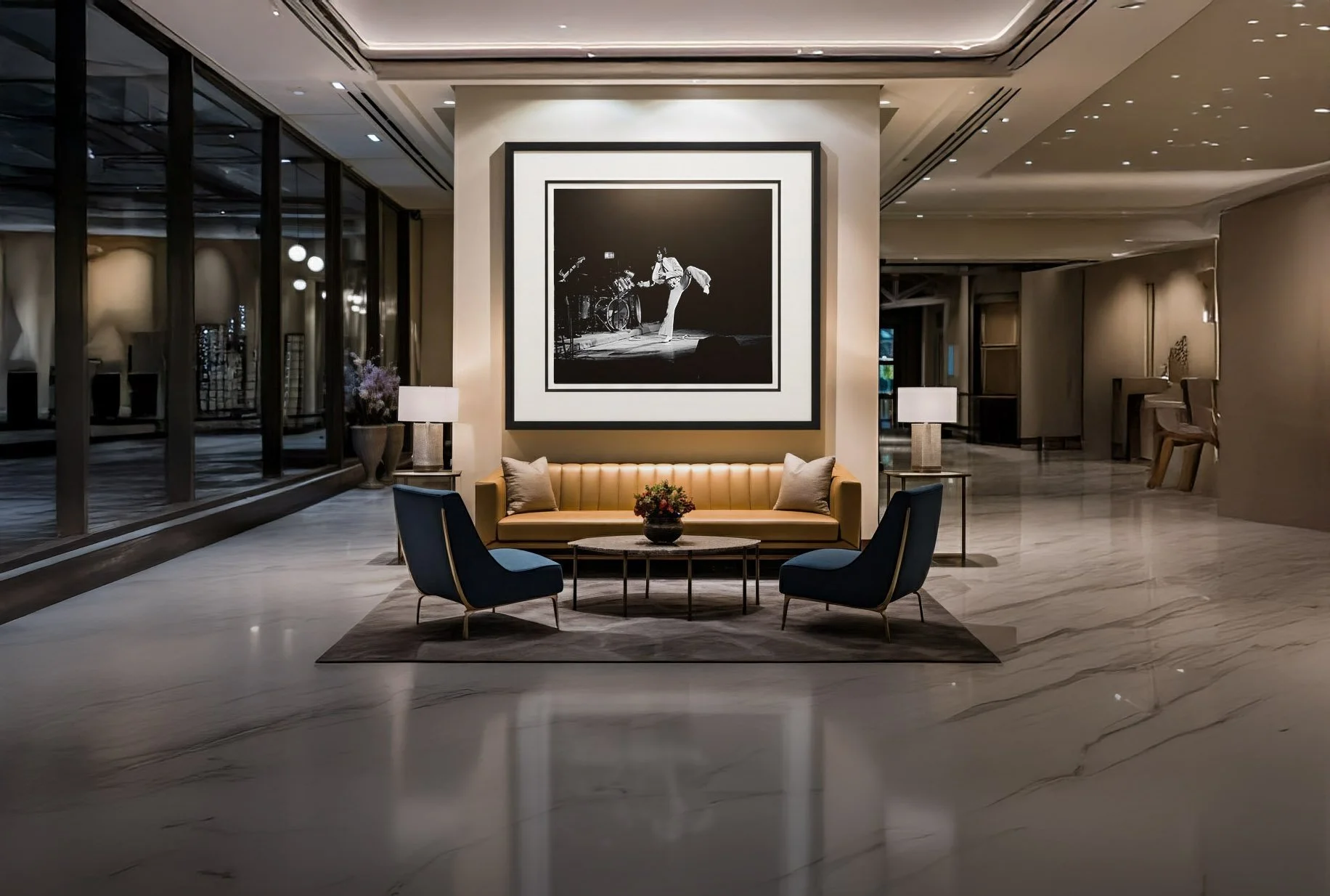 A hotel lobby with a yellow sofa, two blue armchairs, a coffee table with a flower arrangement, and two lamps, with black and white artwork of a person sweeping a bicycle in the background.