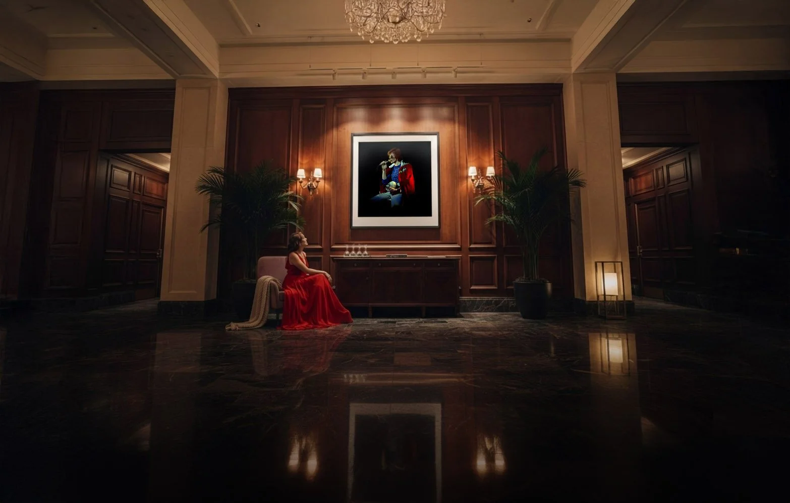 A woman in a red gown sits on a bench in a luxurious hotel lobby, facing a large framed portrait of a singer holding a microphone. The lobby features dark wood paneling, tall potted plants, wall sconces, and a chandelier, with reflections on a polished marble floor.