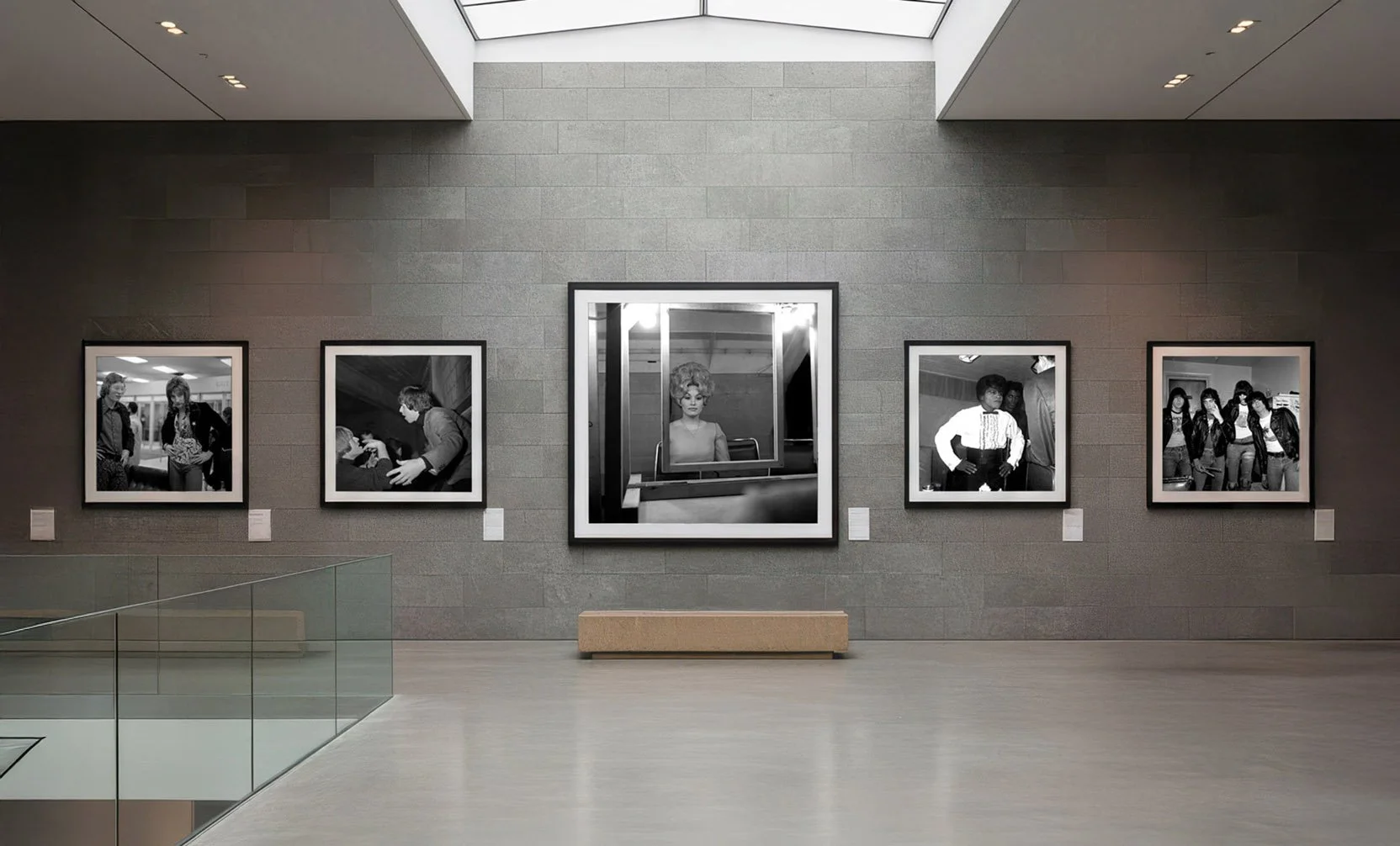 Gallery wall with five black and white photographs, centered by a mirror reflecting a woman, in an art museum or gallery space.