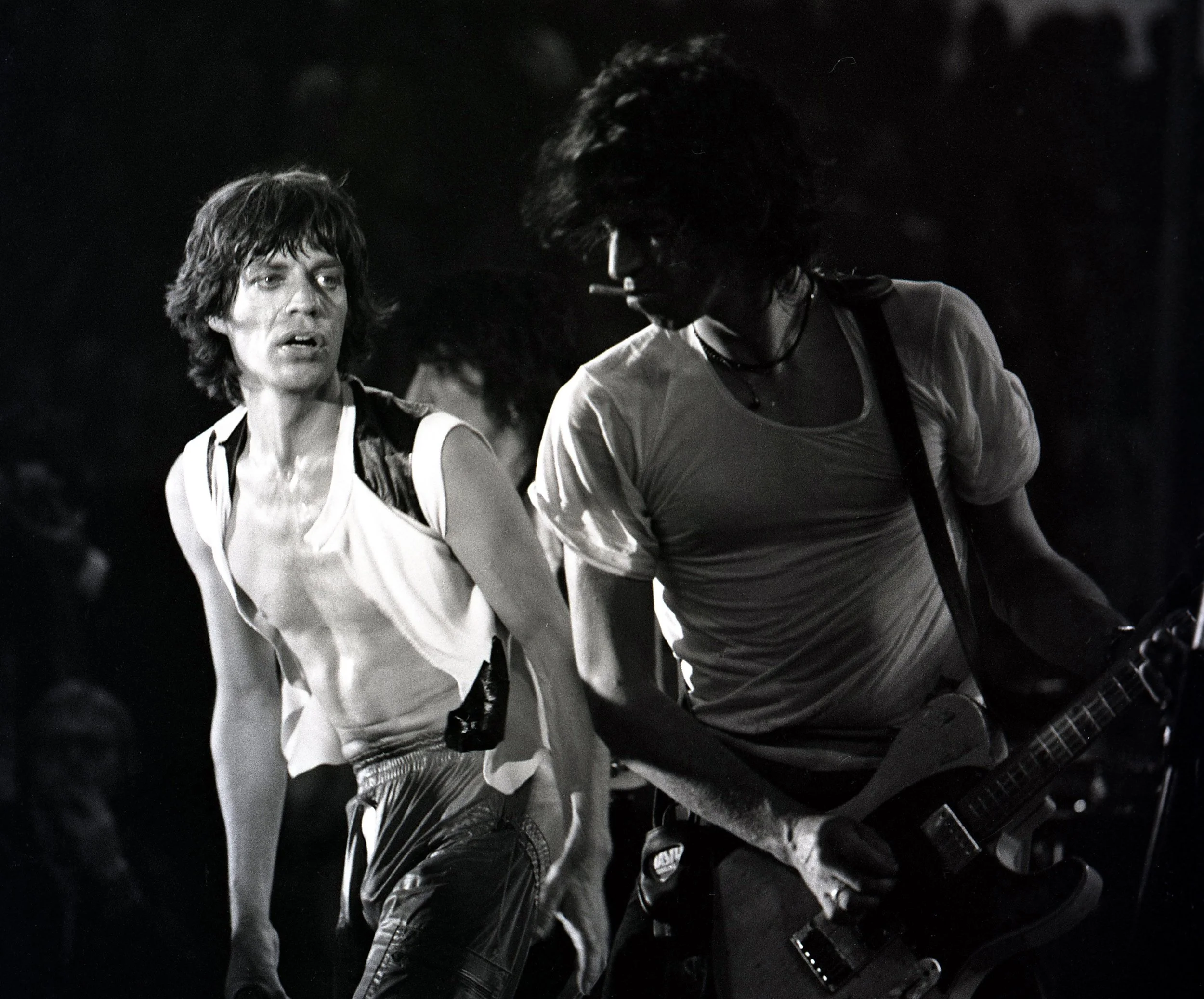 Black and white photo of two men, one with a cigarette in his mouth, wearing a tank top, and the other with a jacket, both appearing to be performing or rehearsing with musical instruments.