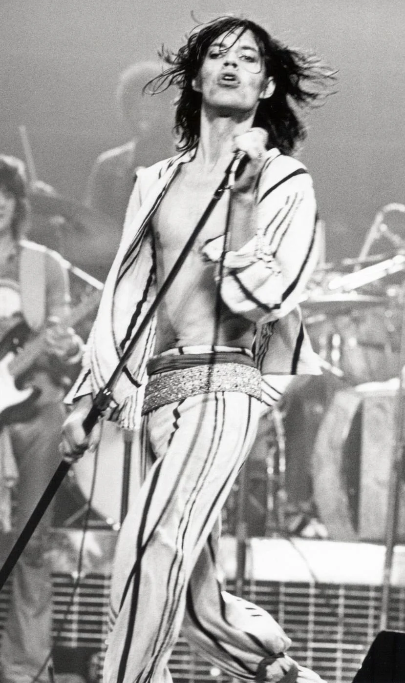 Black and white photo of a woman singing passionately on stage with a microphone, her hair flying, wearing striped pants and a matching top, with band members playing instruments in the background.
