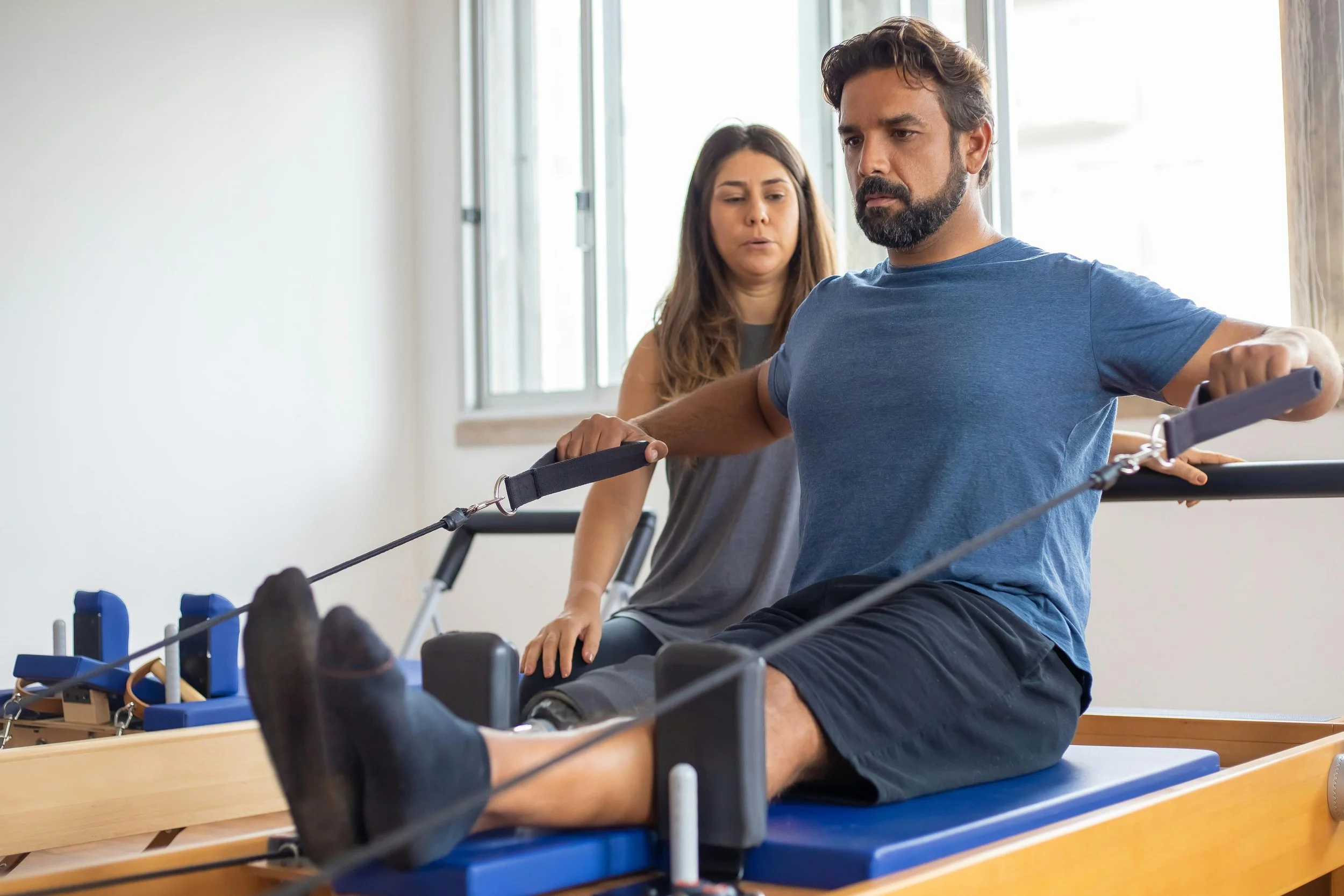 A man doing physical therapy exercises on a reformer Pilates machine with a Pilates teacher assisting him.