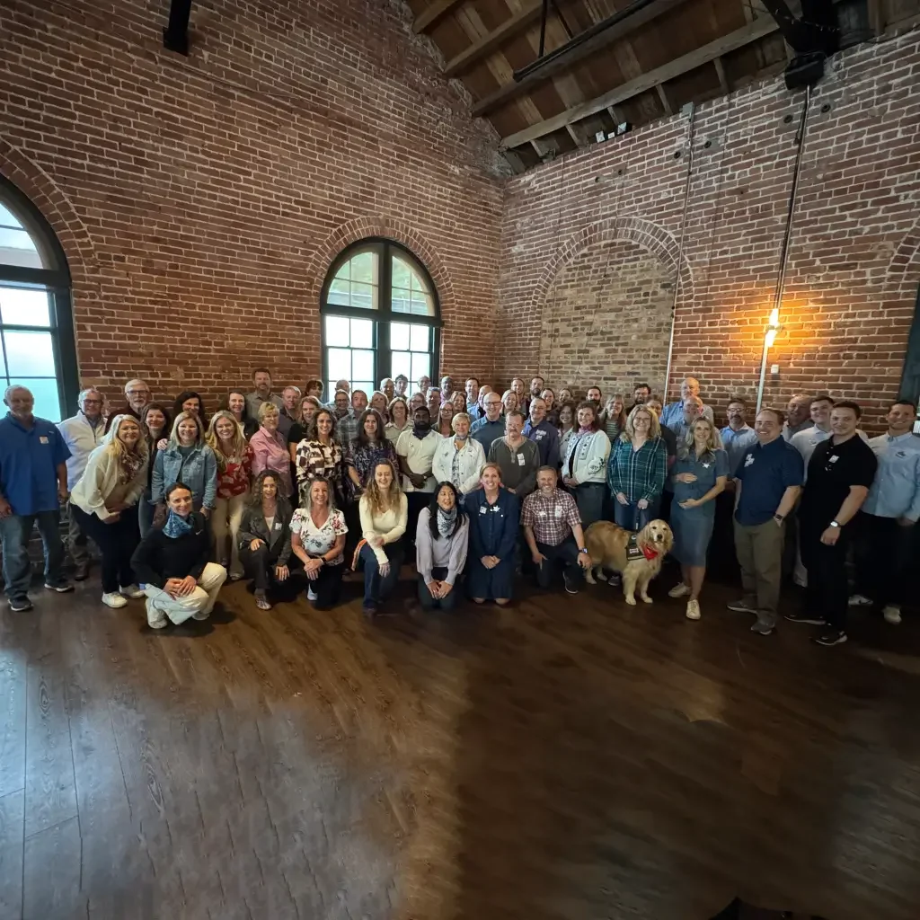 Himformatics Associates- a large group of people gathered in a spacious room with exposed brick walls and wooden ceiling beams, posing for a group photo. There are two large arched windows in the background and a dog near the front.