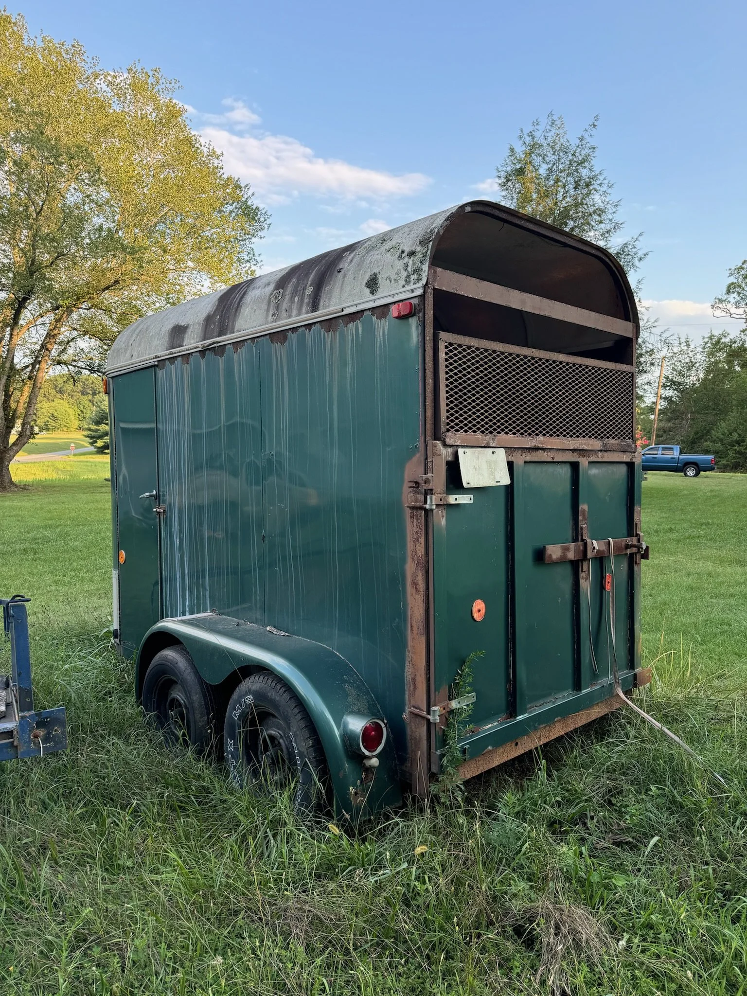A green horse trailer with rust and moss, parked on grass near trees, with a blue pickup truck in the background.