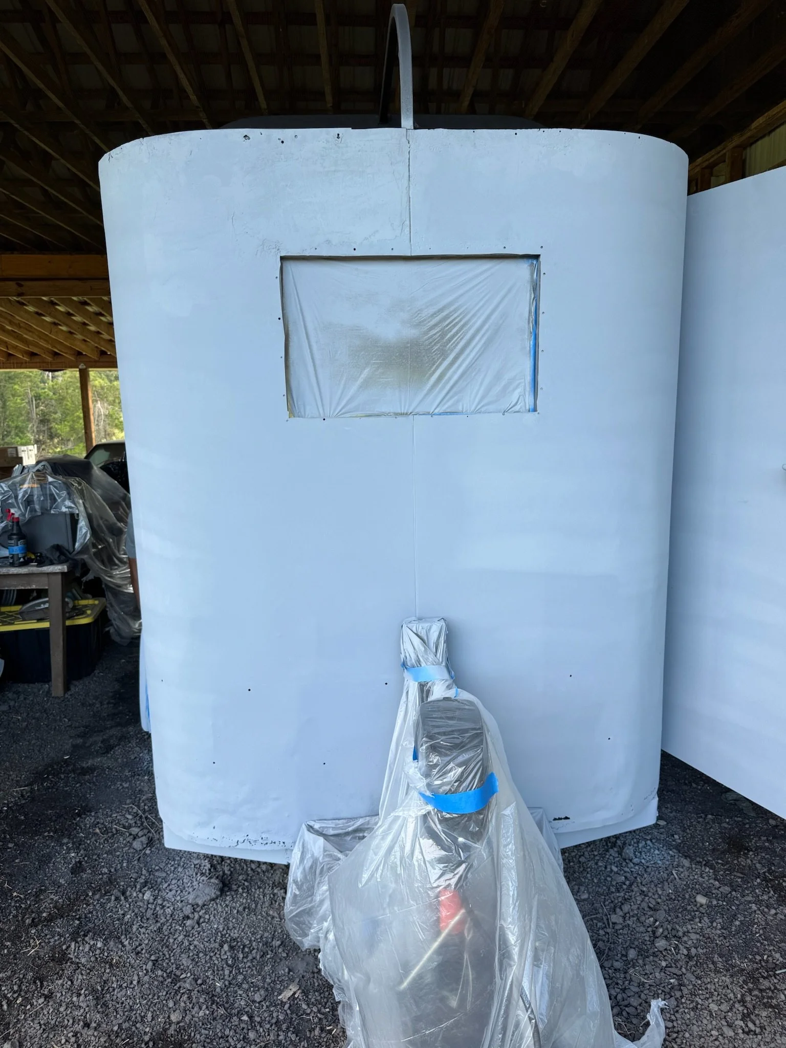 A large white object covered with a plastic sheet, possibly in a workshop or storage area, with a window cutout covered with plastic. There are tools and objects visible on the left side, and the ground is gravel.