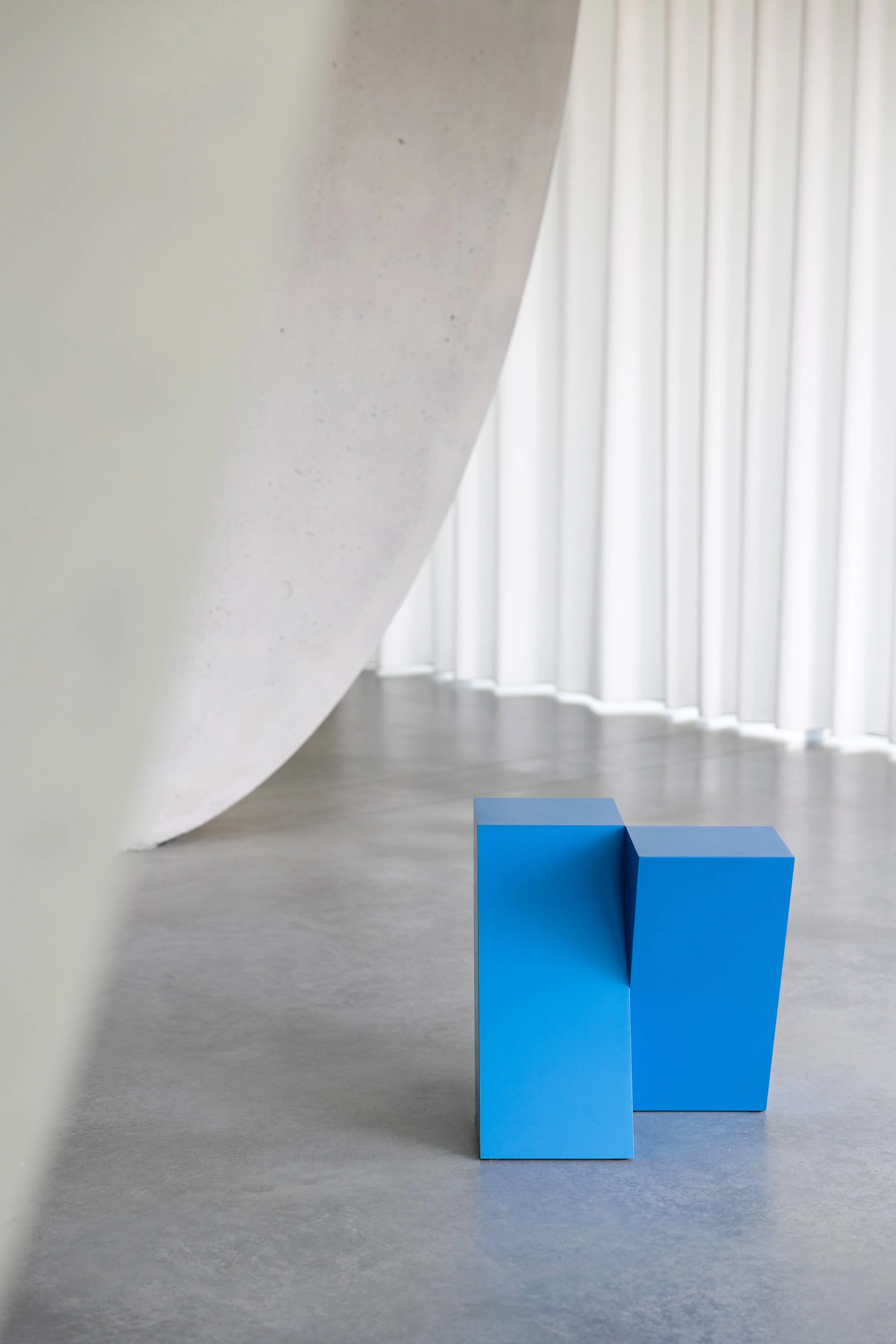 Contemporary art gallery with a blue geometric sculpture on the gray floor, white walls, and tall white curtains in the background.