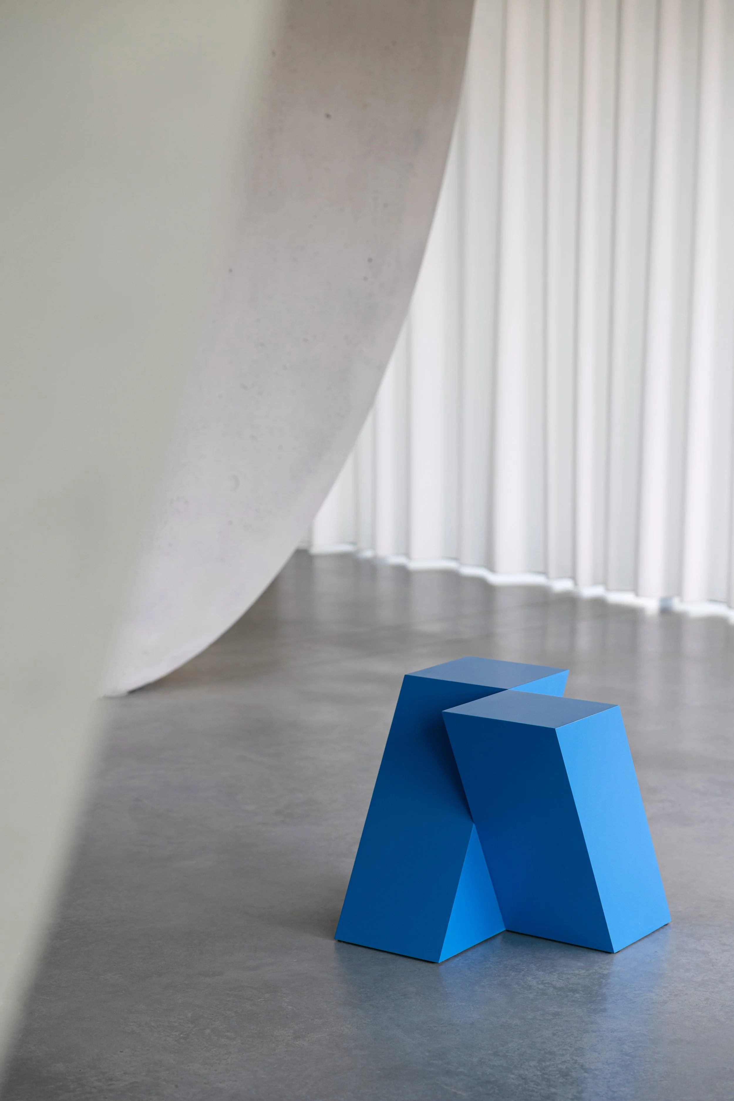 Minimalist art gallery with a blue geometric sculpture on a gray floor, large white curtains in the background, and a curved concrete wall on the left.