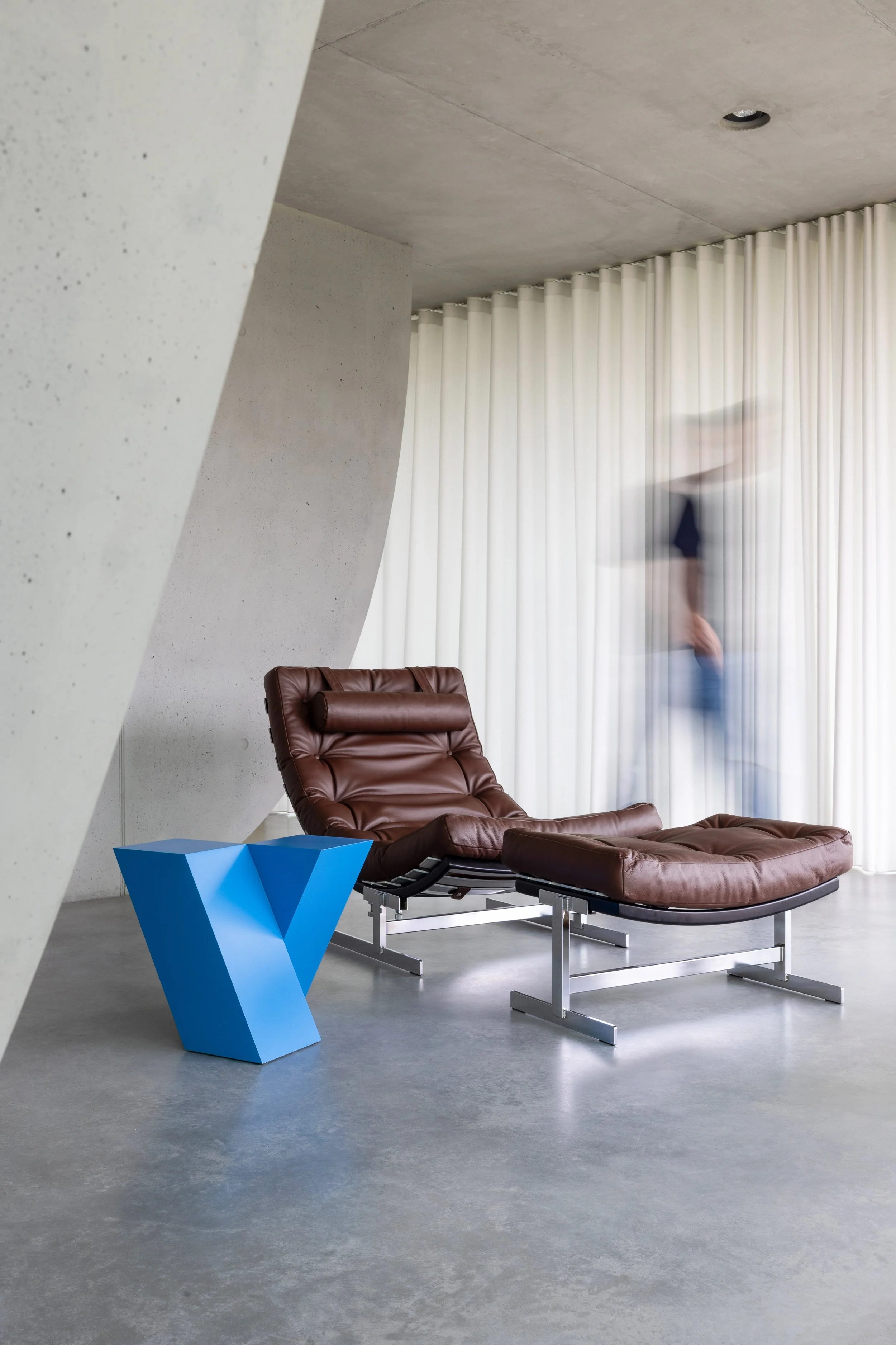 Modern interior room with a brown leather lounge chair, a blue geometric side table, white curtains, a concrete wall, and a blurred person walking in the background.