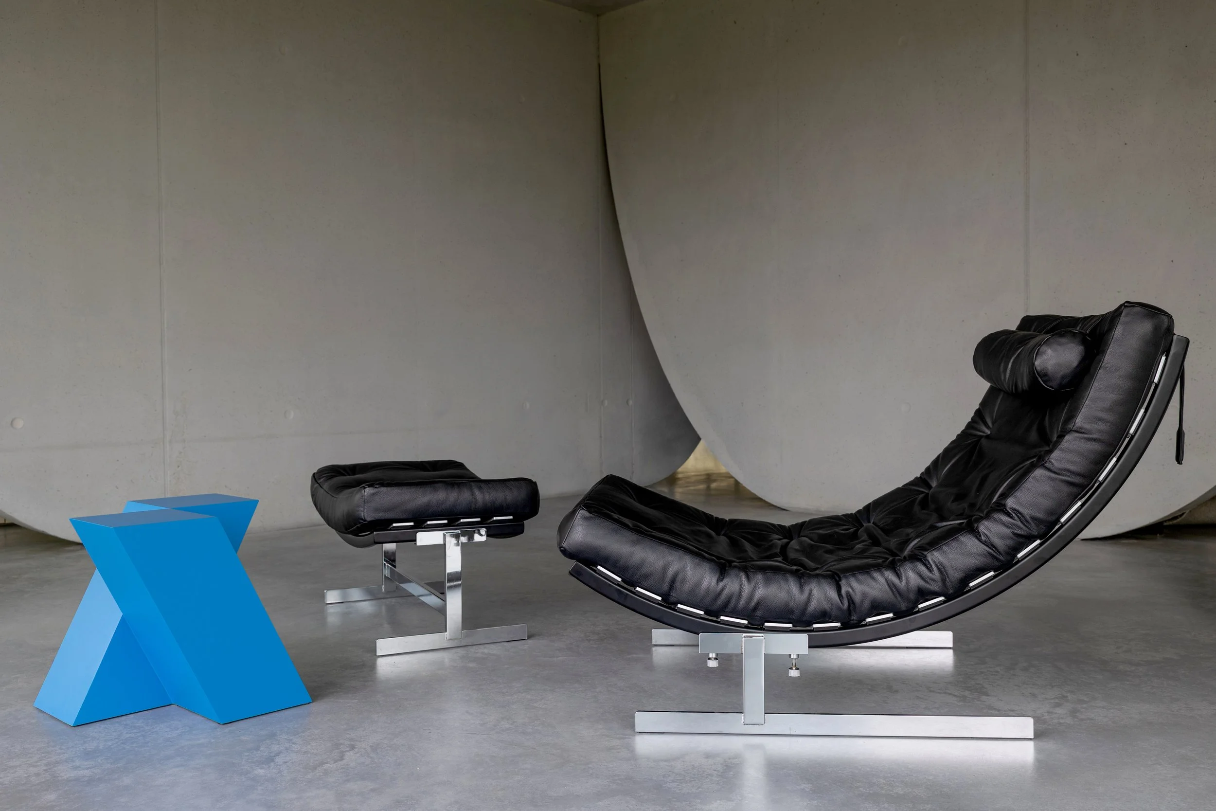 Modern black leather lounge chair and matching ottoman in a room with a concrete floor and gray wall. A blue geometric side table is in front of the chair.