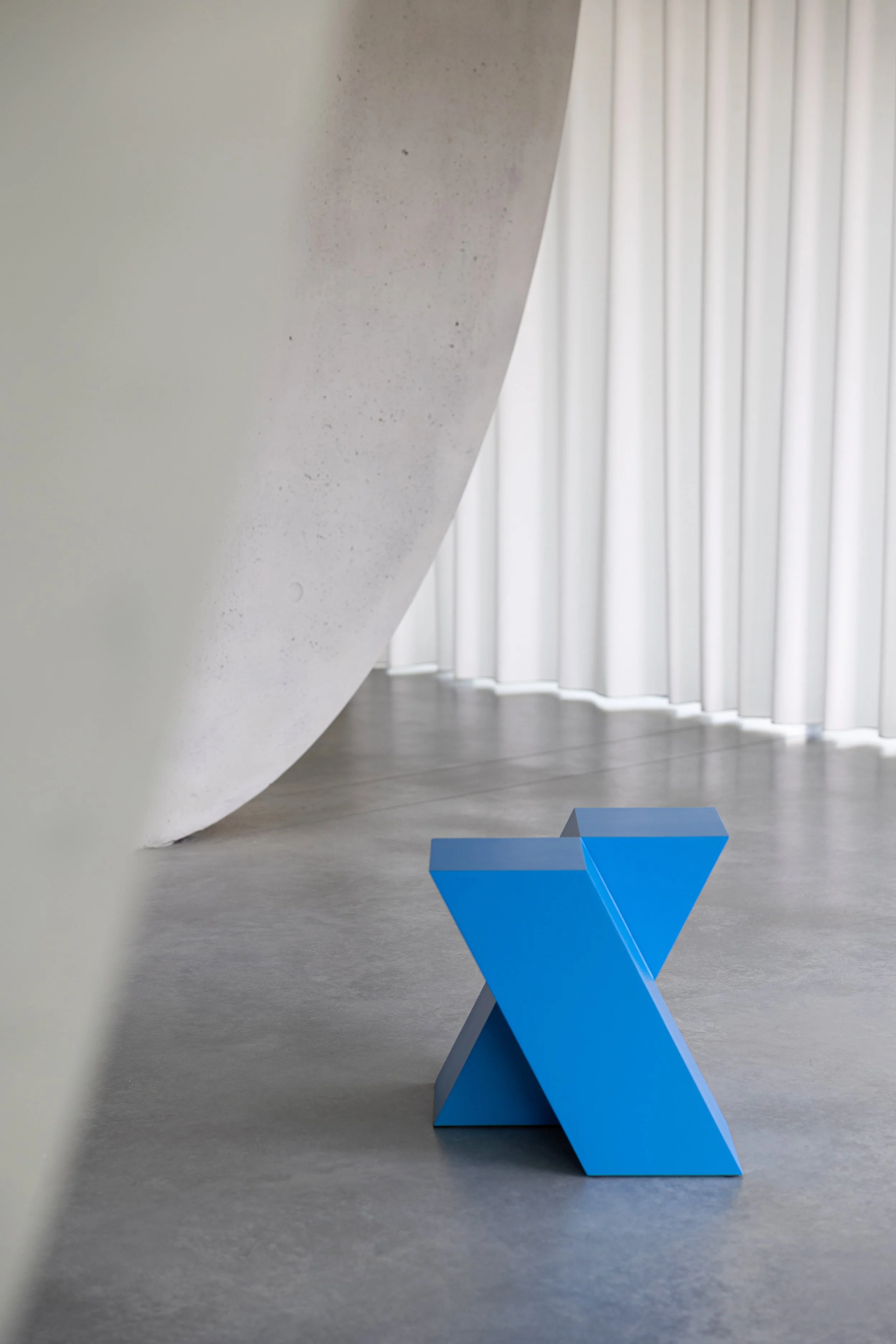 Modern interior space with a blue geometric sculpture on a gray floor next to a white curved wall and white curtains in the background.