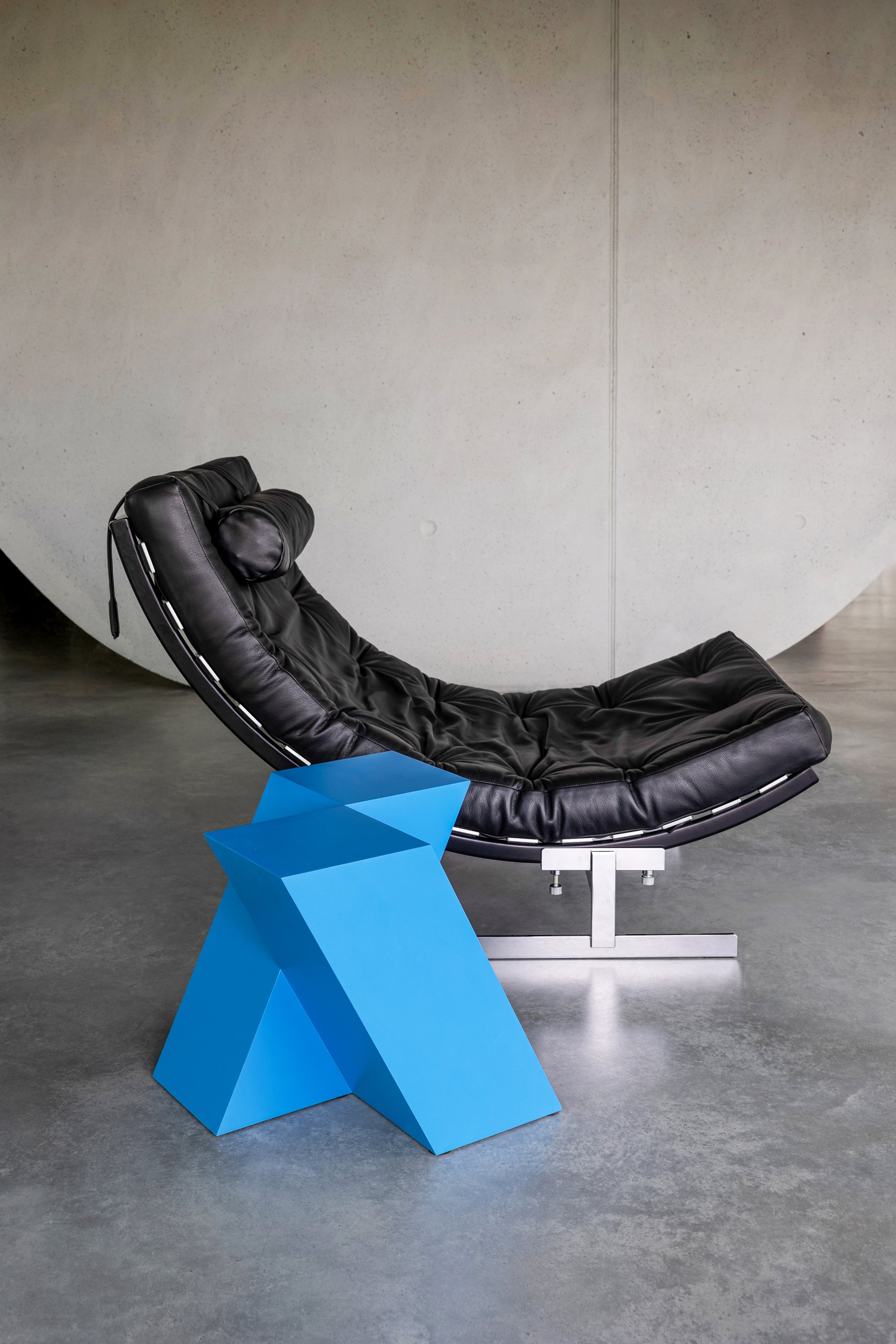 Modern black lounge chair with a pillow on a blue geometric side table placed on a gray concrete floor against a plain light gray wall.