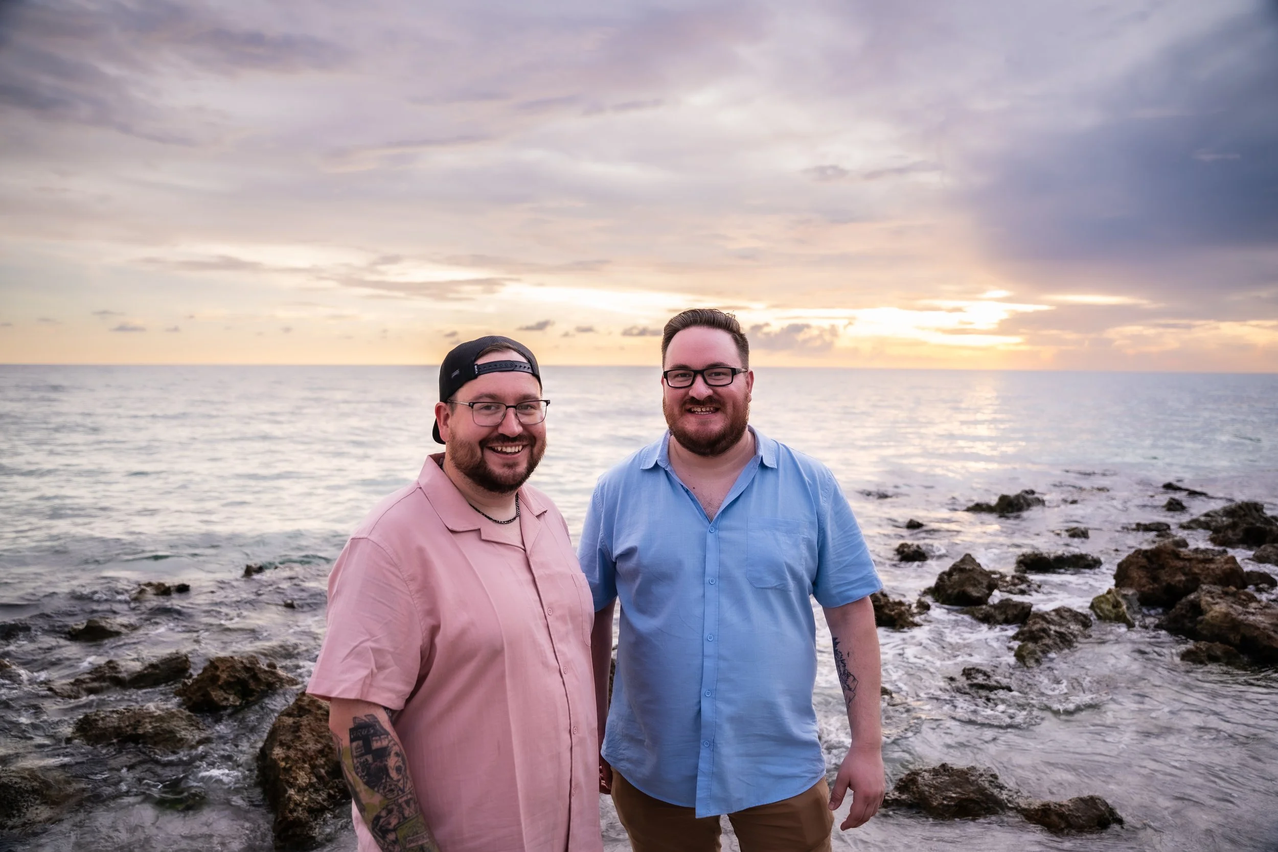 Two men standing on rocks by the ocean at sunset, smiling at the camera.