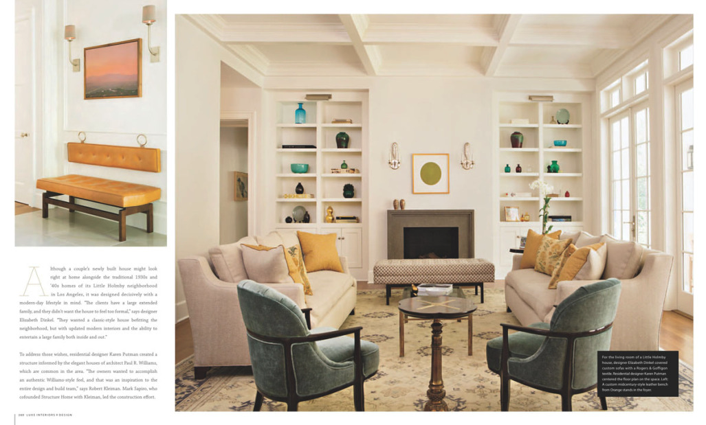 Living room with two beige sofas, two green armchairs, a fireplace, built-in bookshelves, large windows, and decorative objects. Small side table and a rectangular ottoman in front of the fireplace.