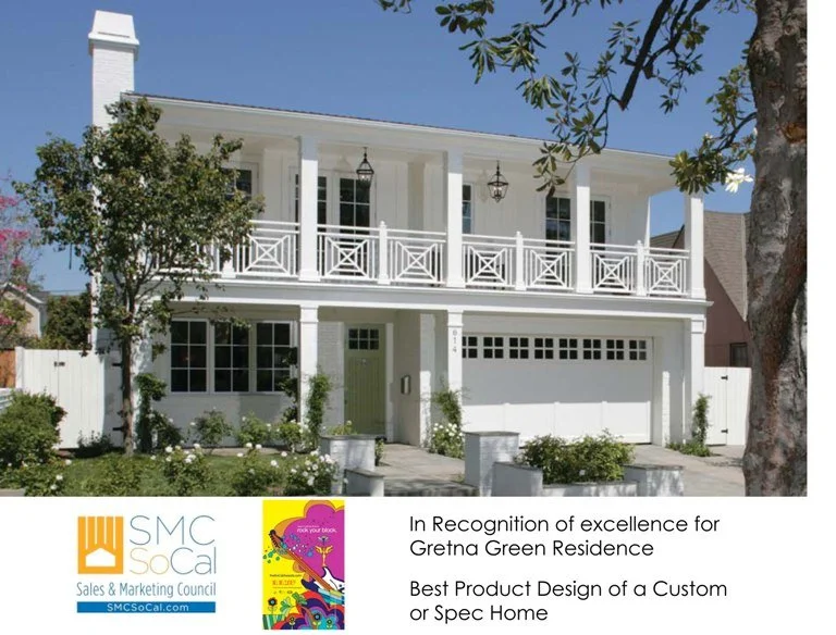 A two-story white house with a front porch, decorative railing, and hanging lanterns, surrounded by trees and shrubs.