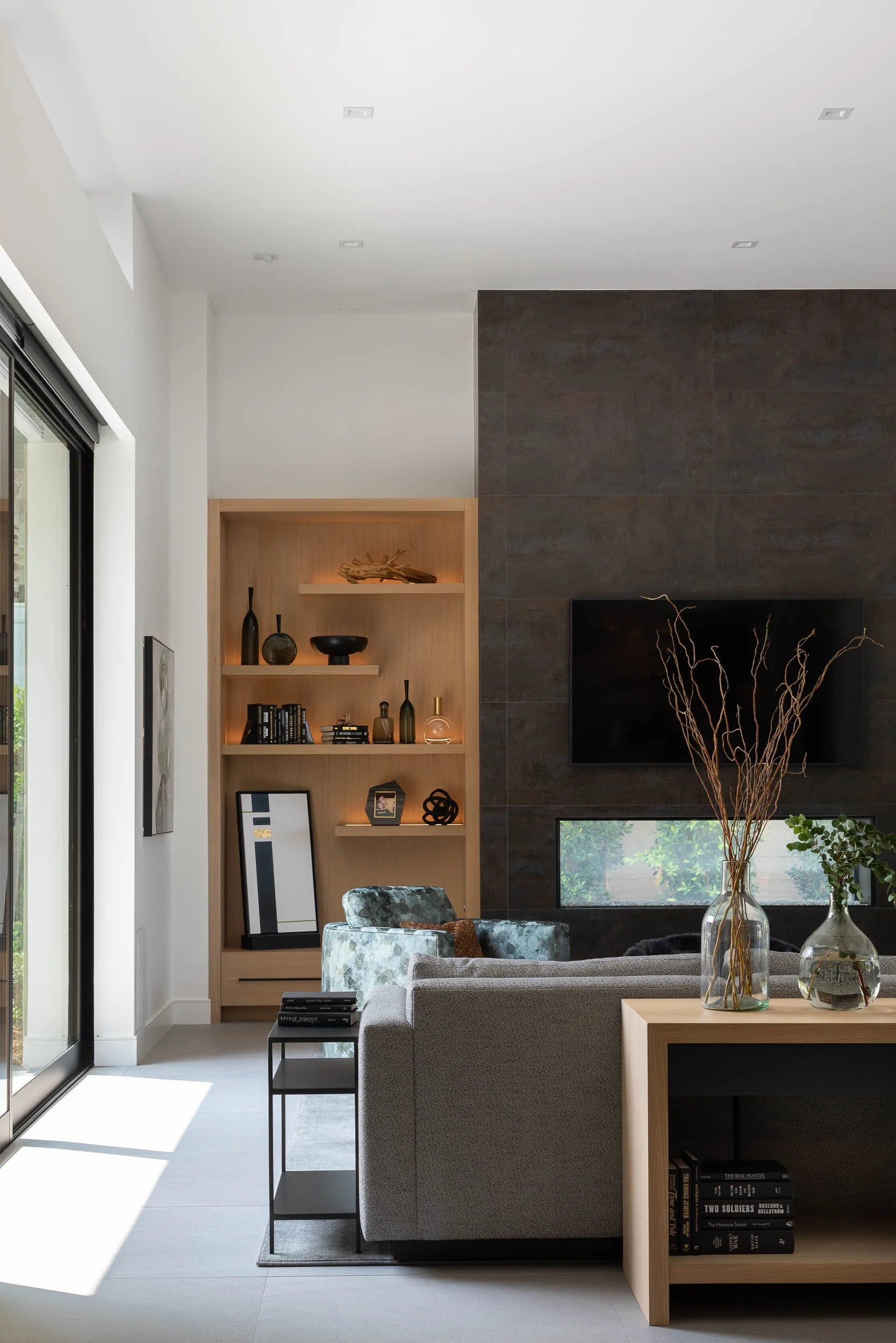 Modern living room with a gray sofa, black side table with books, a built-in wooden bookshelf with decorative items, a large flat-screen TV, and vases with branches and greenery, lit by sunlight through sliding glass doors.