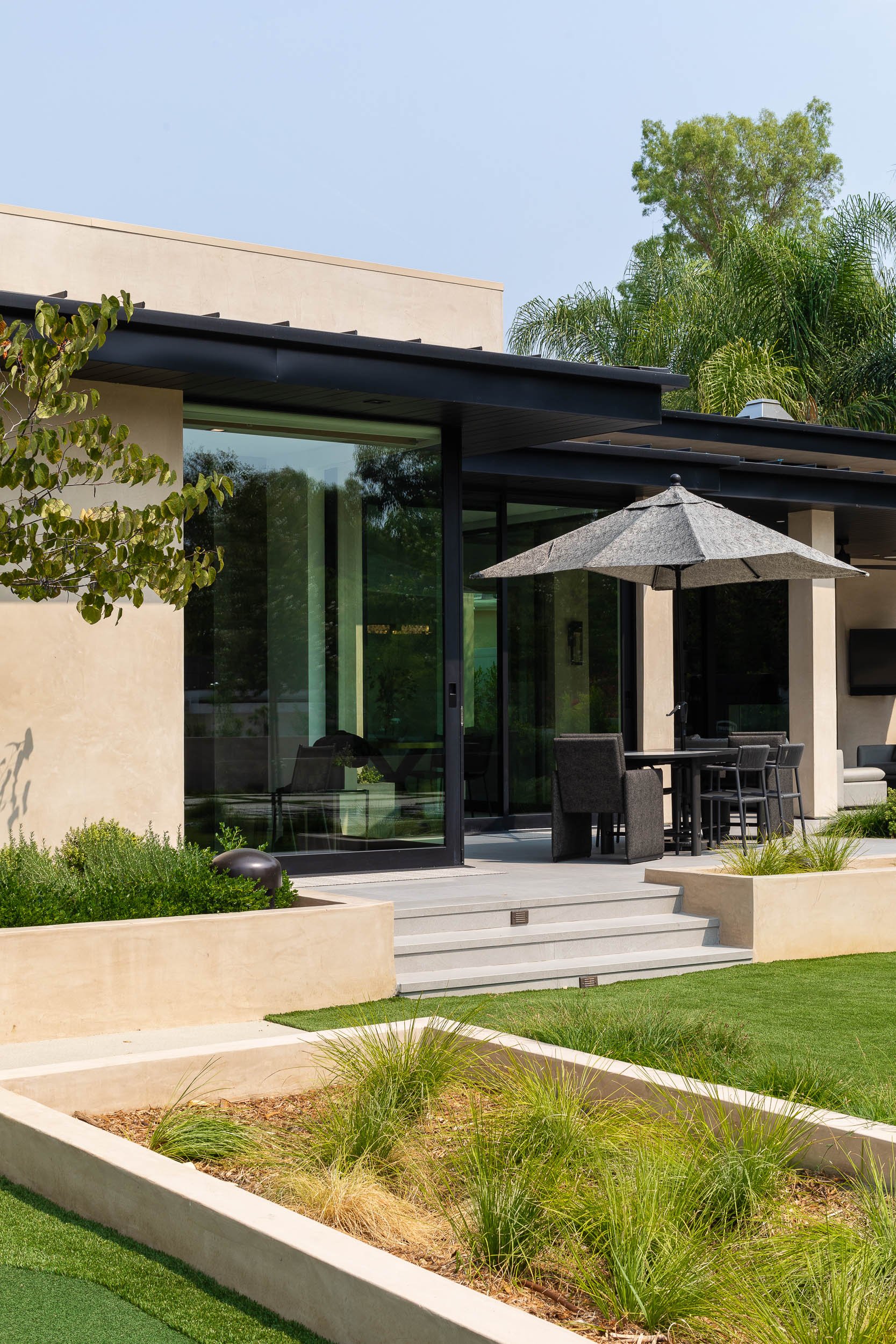 Modern house with large glass windows, patio furniture with an umbrella, and a green lawn with landscaped plants under a clear sky.