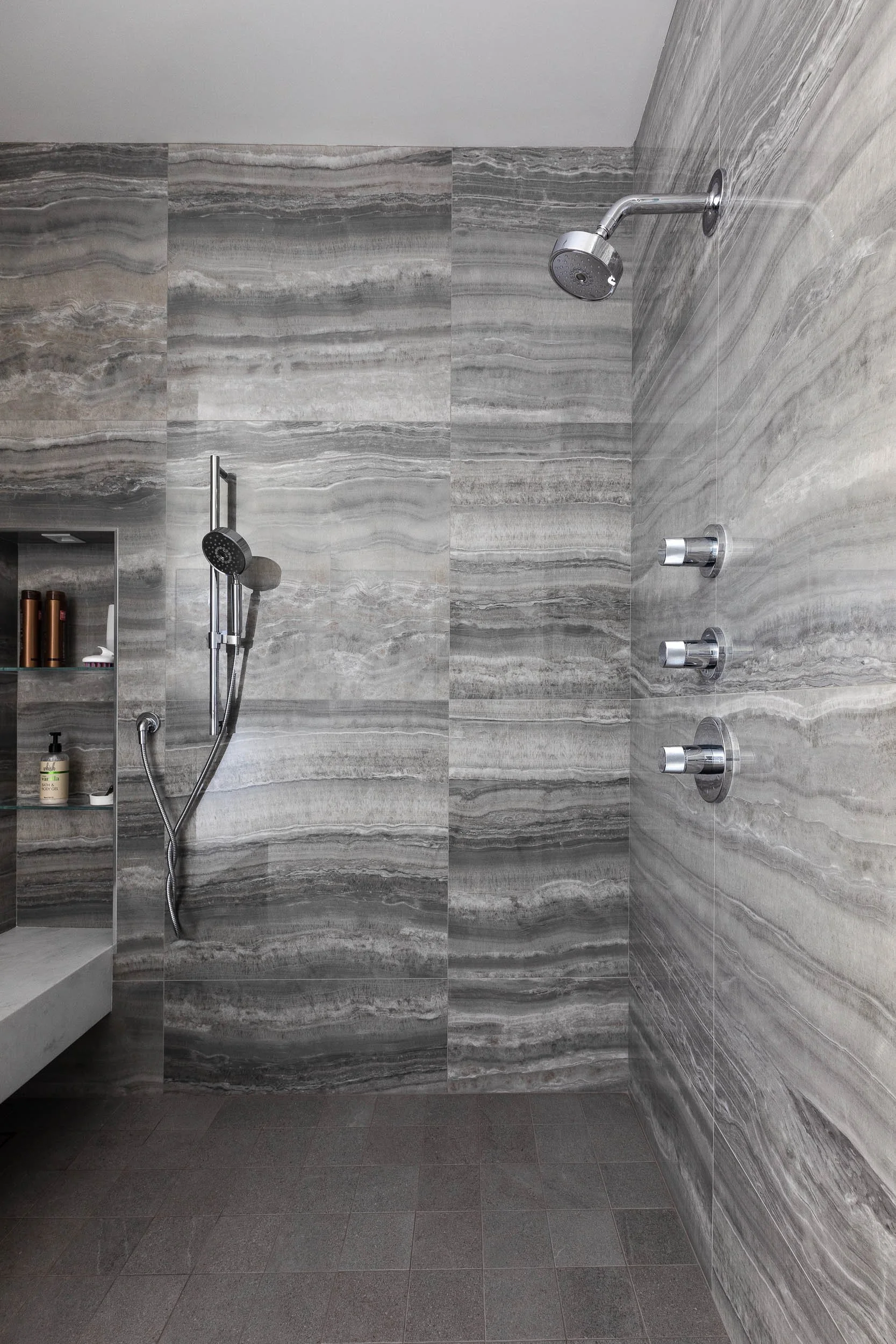 A modern walk-in shower with gray marble tiles on the walls and dark gray tile on the floor. Features include a fixed shower head at the top, a handheld shower head, and two adjustable body sprays.