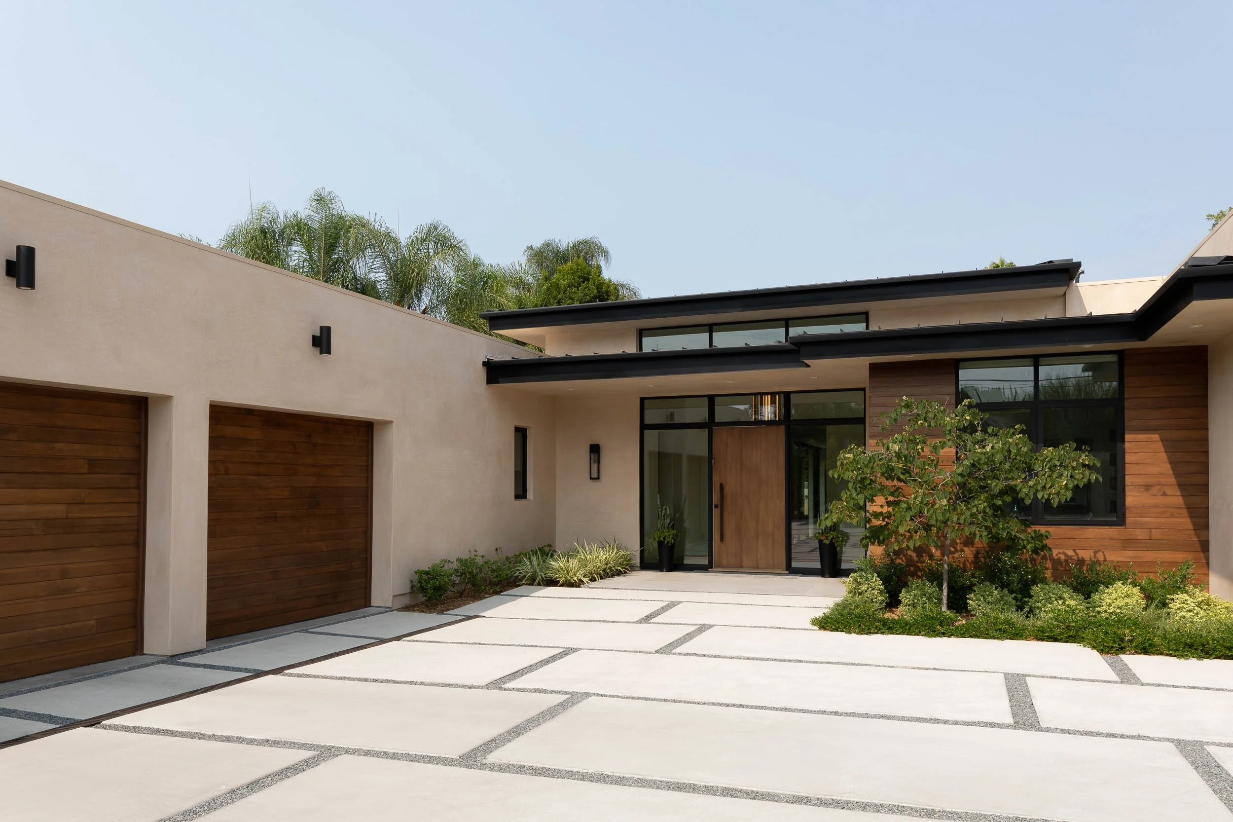 Modern house with a driveway, wooden garage doors, large glass windows, and landscaped front yard with a small tree and shrubs.