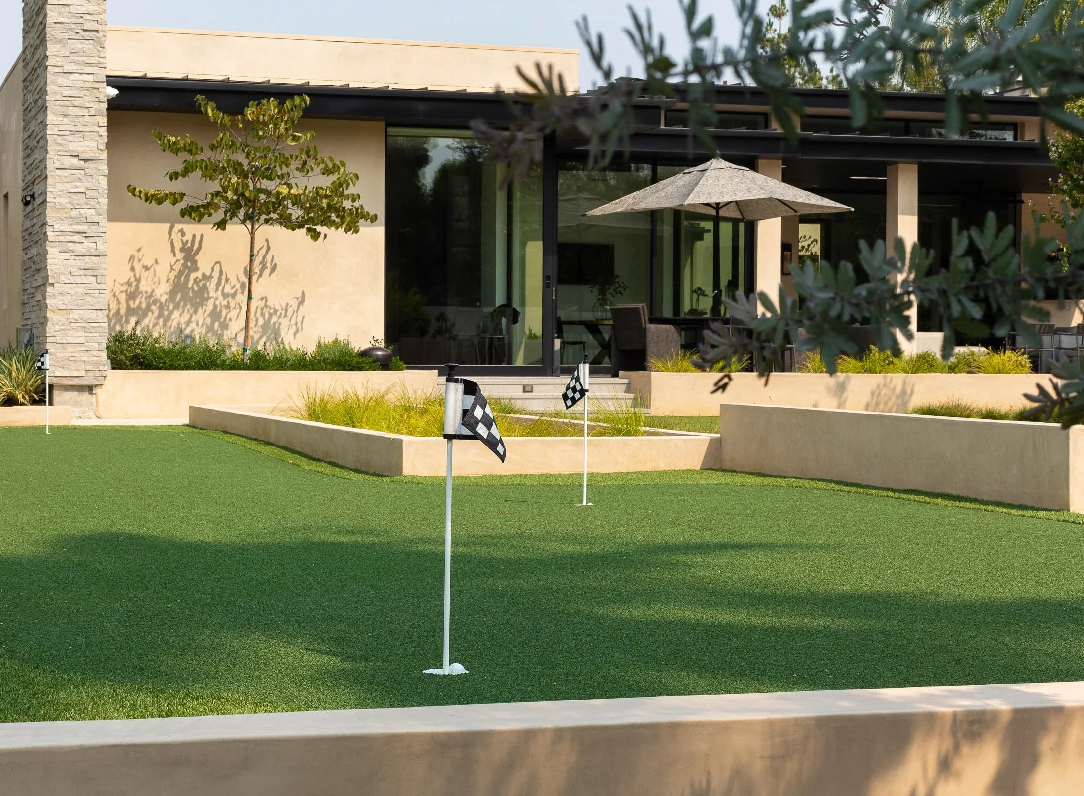 A backyard putting green with two checkered flags, adjacent to a modern home with large glass sliding doors, a patio area with outdoor furniture and a patio umbrella, surrounded by landscaped garden beds and trees.