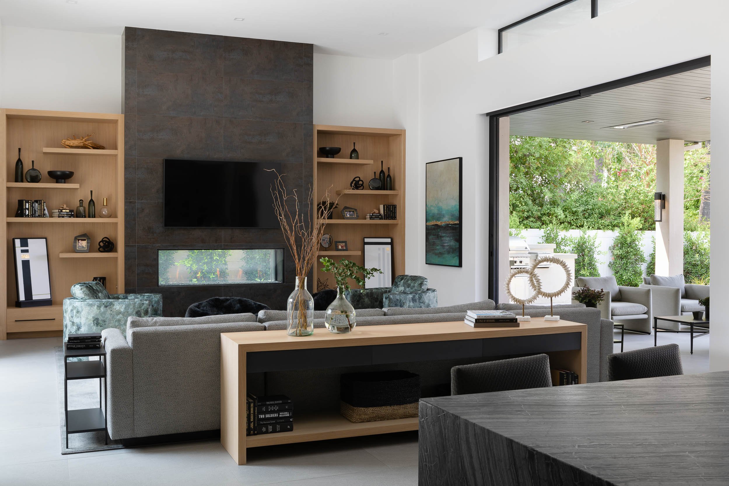 Modern living room with a large sofa, decorative vases, books, a wall-mounted tv, built-in wooden shelves with decor, a sliding door opening to an outdoor patio with chairs, curtains, and greenery.