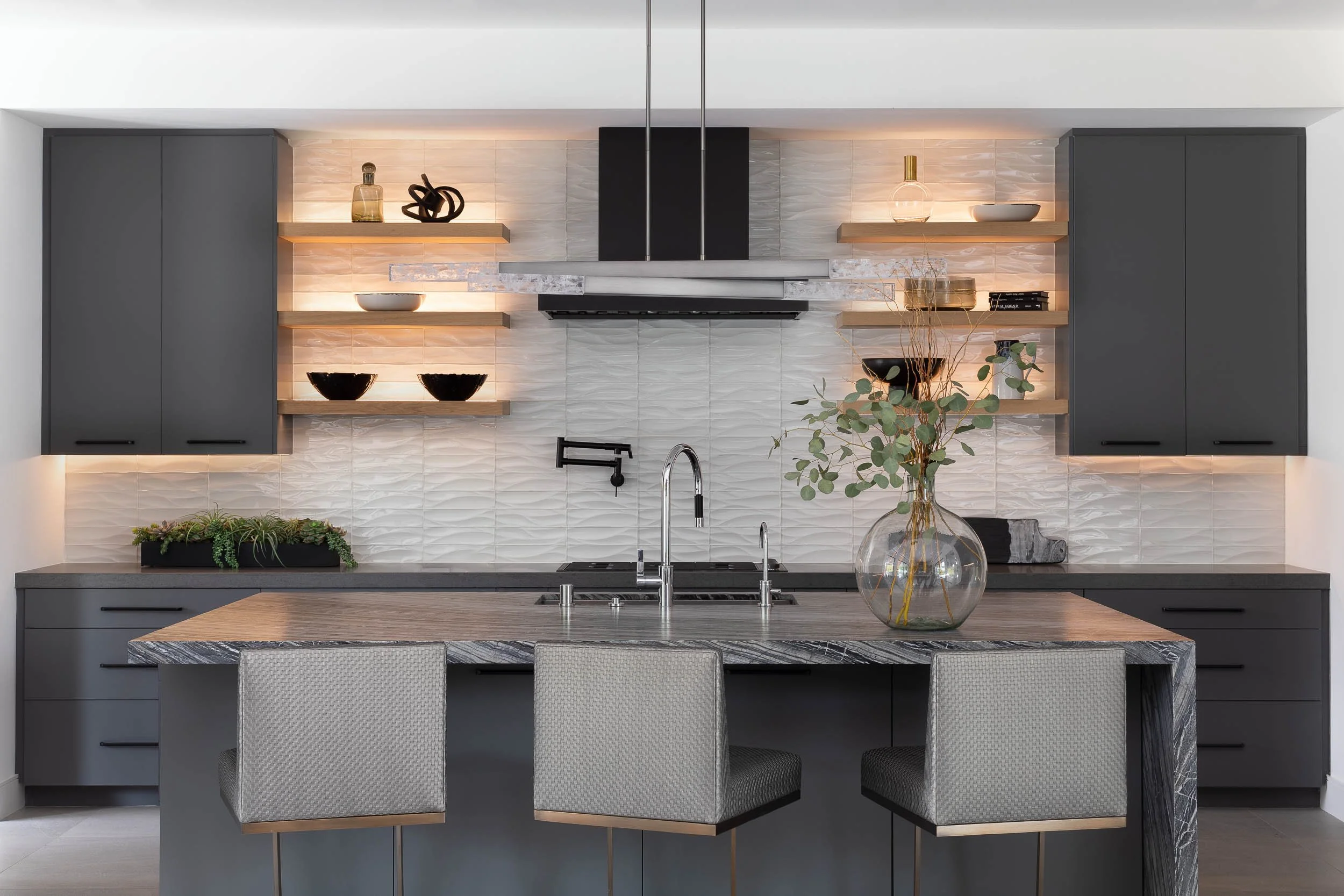 Modern kitchen with dark gray cabinets, wooden open shelves, a marble kitchen island with a vase of green foliage, and decorative items on shelves.