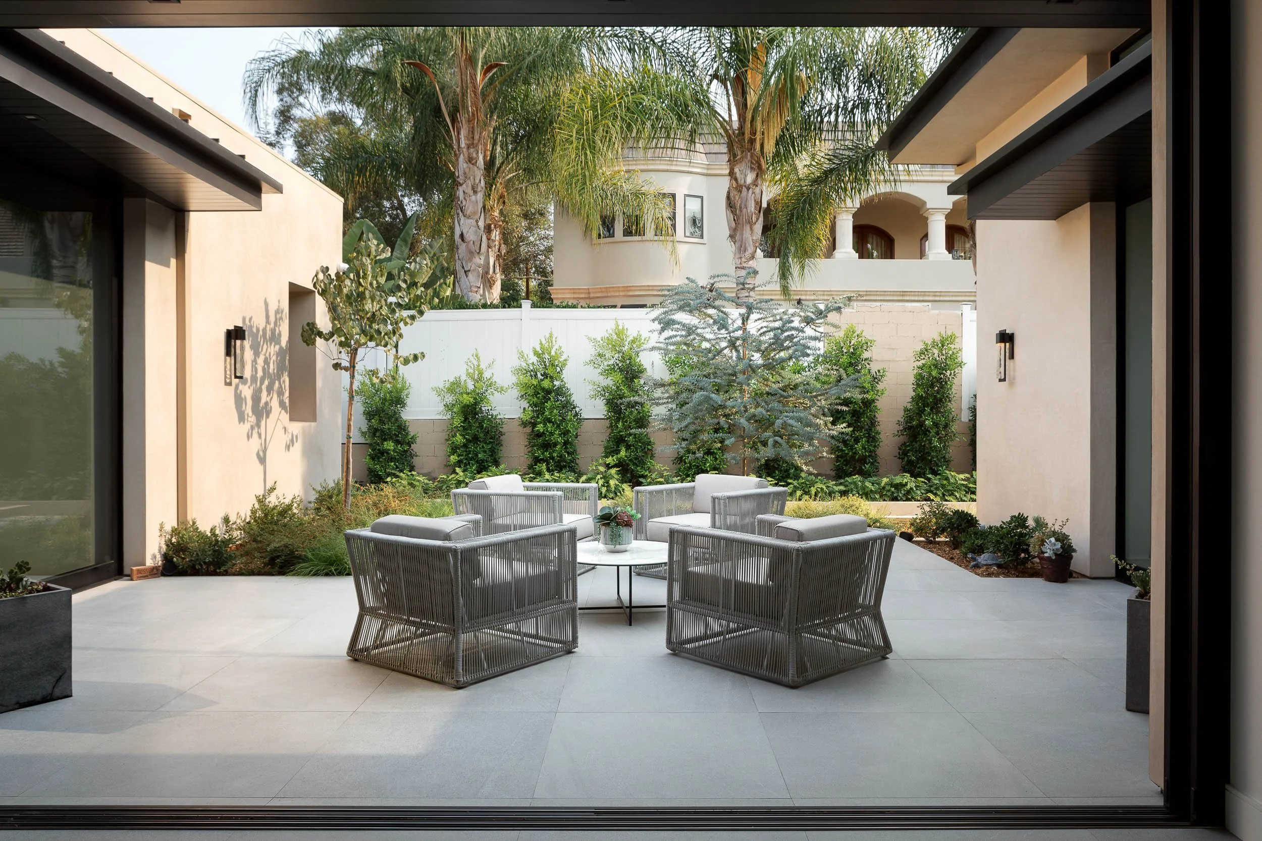 Modern outdoor patio with five gray outdoor chairs arranged around a small round table, surrounded by green plants and trees, with a view of a white house with columns in the background.