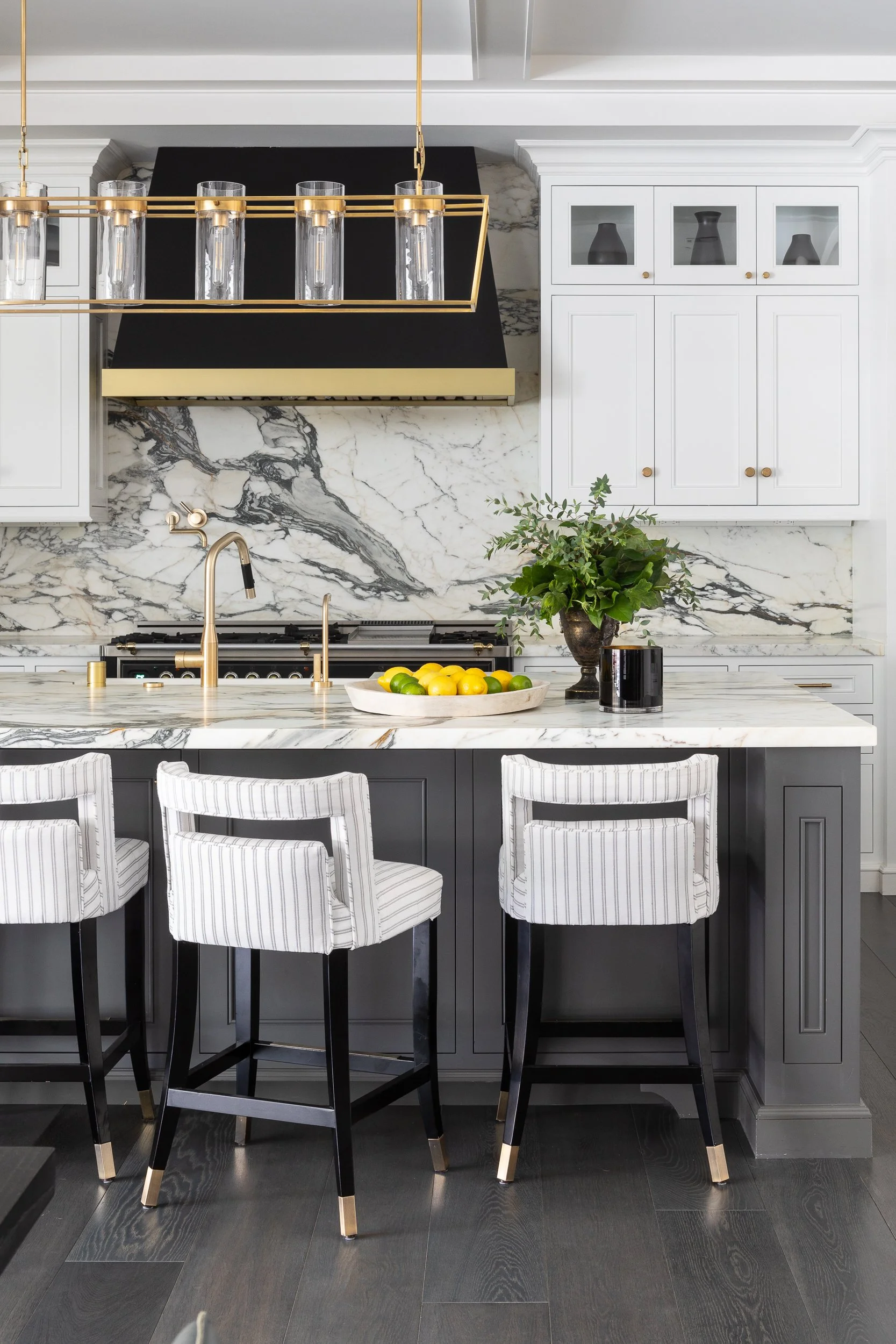 Modern kitchen with marble countertops and backsplash, white cabinetry, black and gold accents, and striped bar stools.