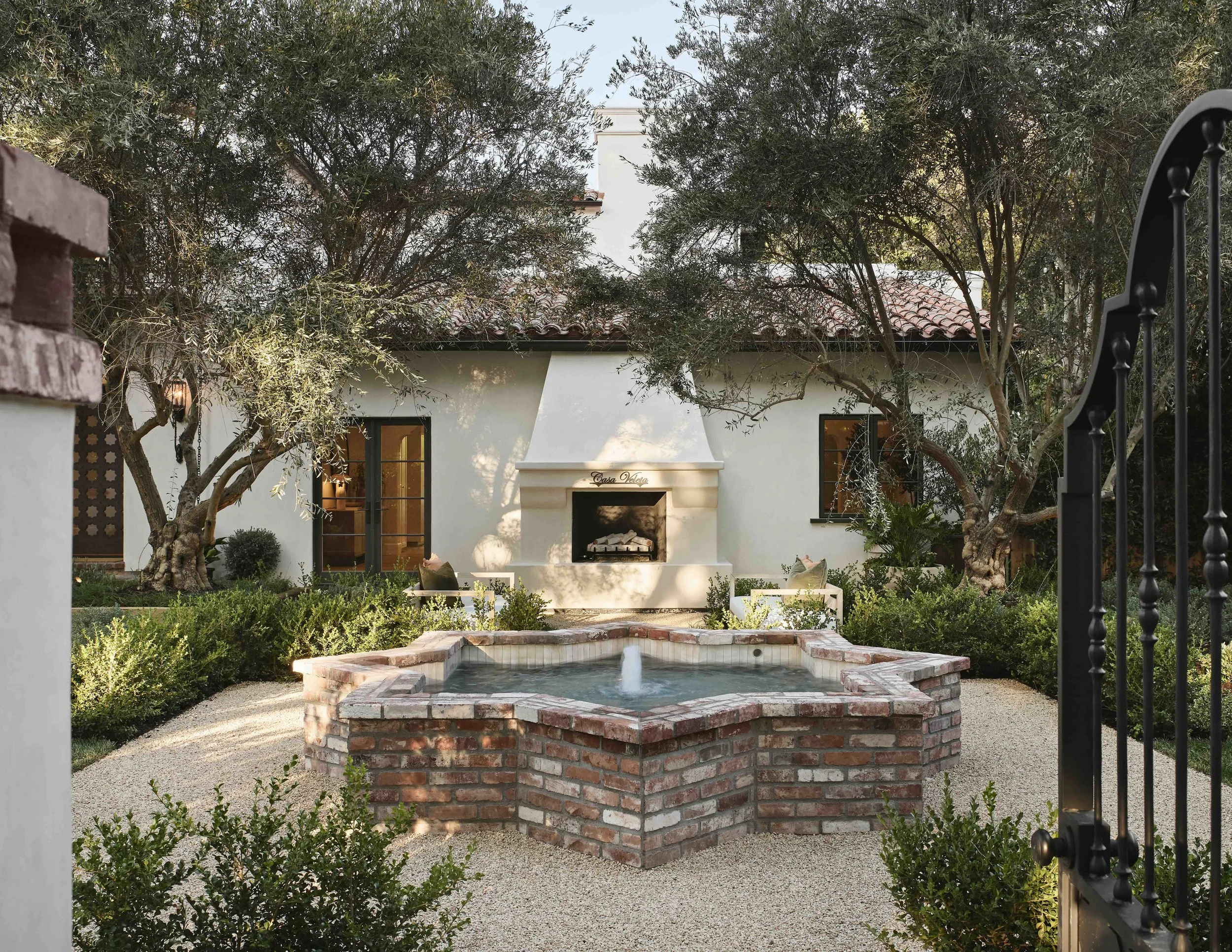 A white house with a fireplace, surrounded by trees and bushes. In front, there's a brick fountain with a small water feature. The view is framed by a black wrought iron gate on the right.