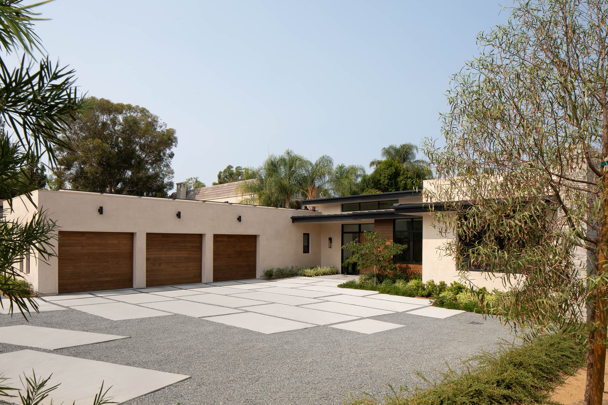Front view of a modern house with three garage doors, concrete pavers in the driveway, and landscaping with trees and shrubs.