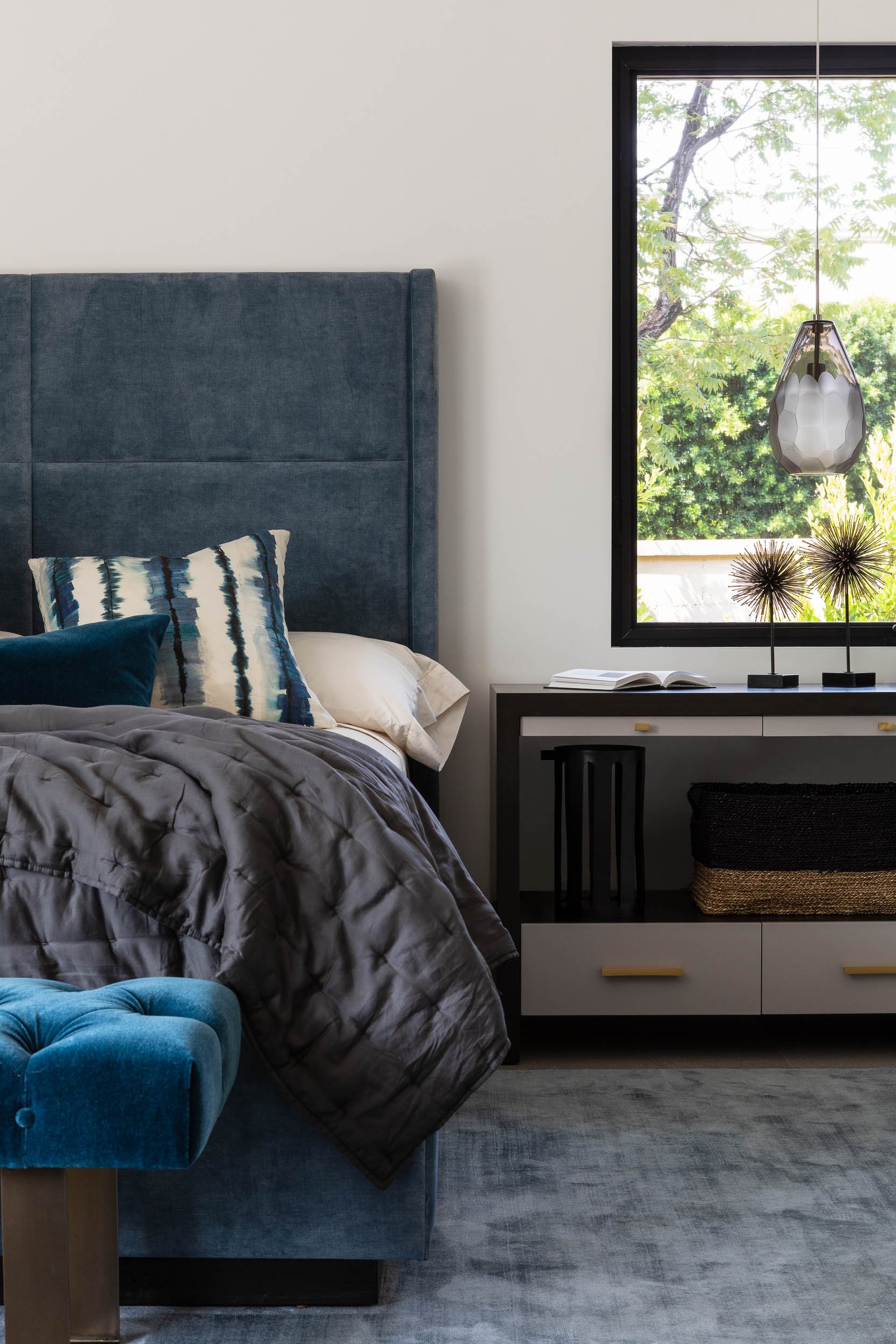 A bedroom with a tall teal upholstered bed with pillows, a gray quilt, and a blue velvet bench. A window with green trees outside, a black-and-white dresser with decorative objects, and a gray rug.