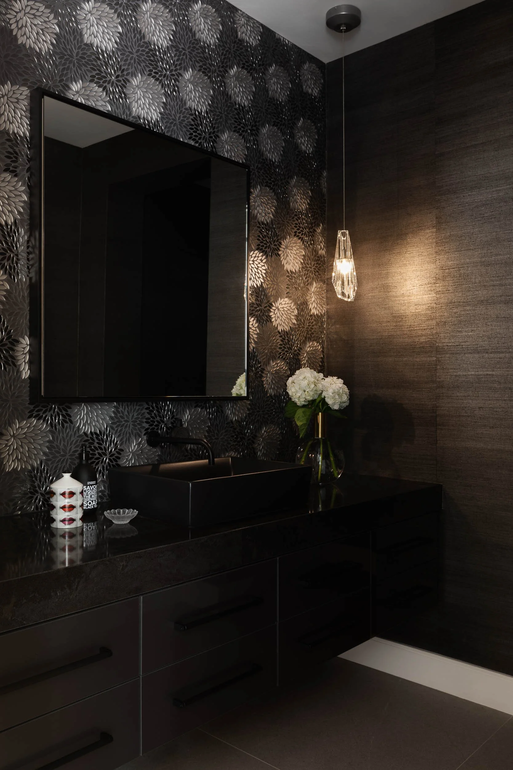 A modern bathroom with a black vanity, rectangular black basin, and a large mirror. There are decorative bottles on the counter, a glass vase with white flowers, and a hanging pendant light. The walls have dark-colored textured and patterned wallpape